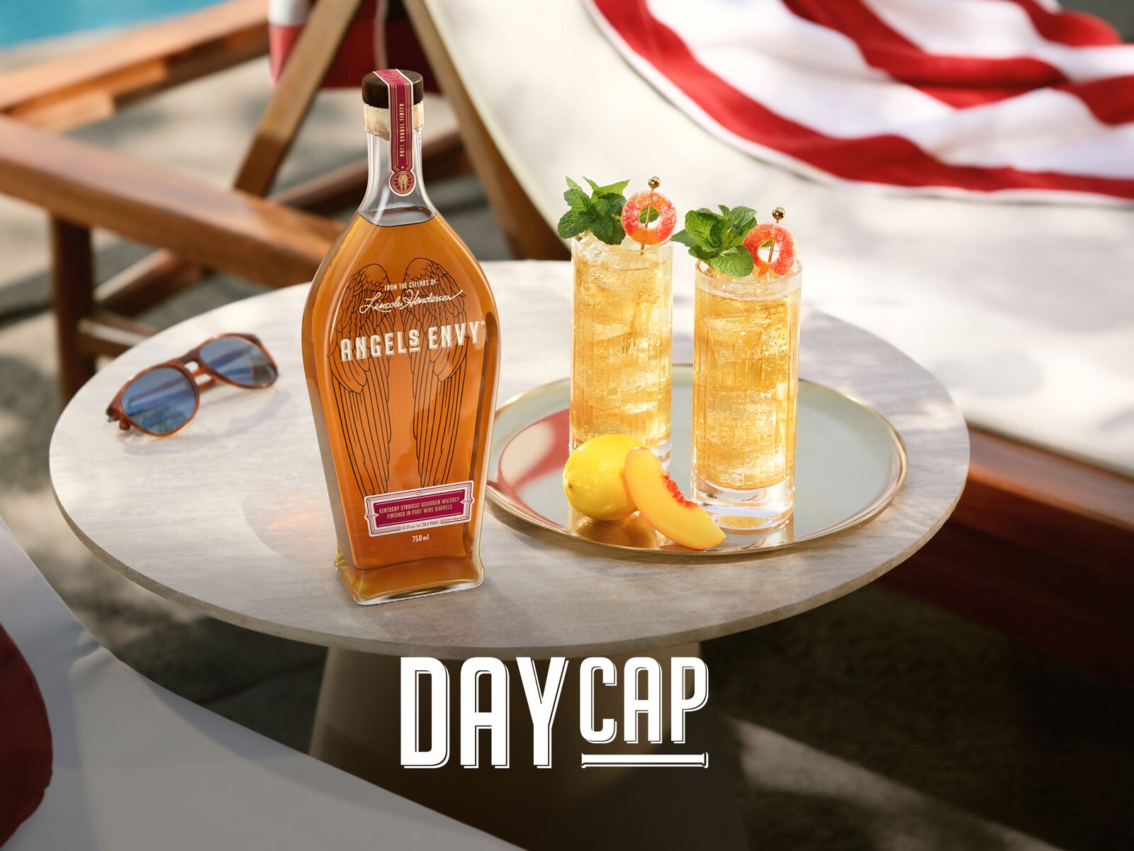 A bottle of Angel’s Envy bourbon with two golden Daycap cocktails on a sunlit poolside table, styled with fresh fruit and mint, on a relaxing summer day.