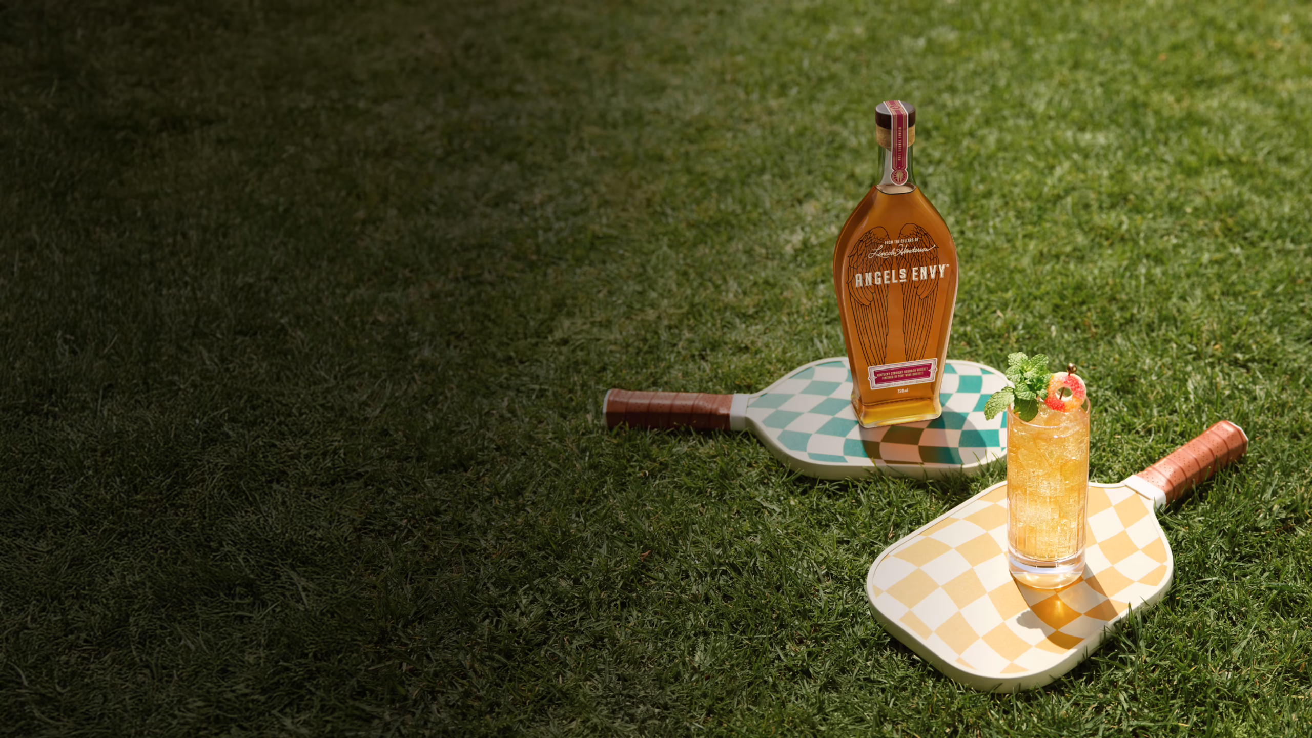 An icy Angel’s Envy Peach Smash cocktail garnished with mint and sour candy. It’s served in a highball glass on top of a patterned, pickleball paddle with a bottle of Angel’s Envy bourbon behind it.