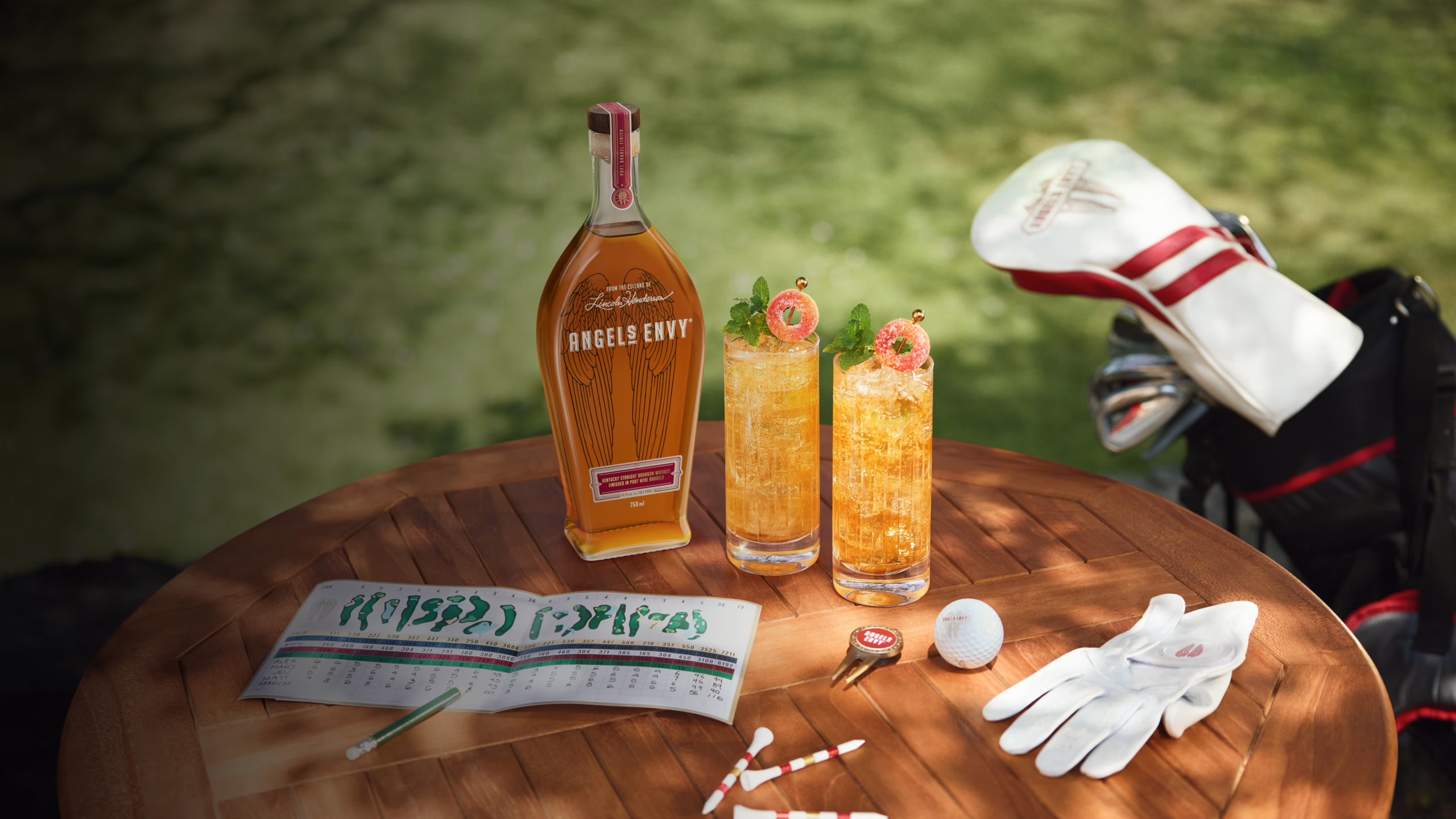 A bottle of bourbon and two icy Daycap cocktails on a outdoor wooden table, surrounded by Angel’s Envy branded golf equipment in warm afternoon light.