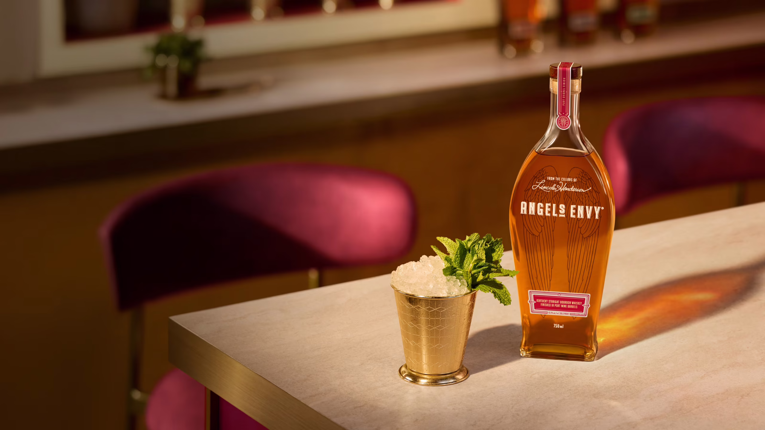 Set in a warmly-lit modern bar with ruby chairs and white tabletop, a bottle of Angel’s Envy 2026 Cask Strength Bourbon is presented alongside an iced Mint Julep cocktail in a golden signature Angel’s Envy Mint Julep cup.