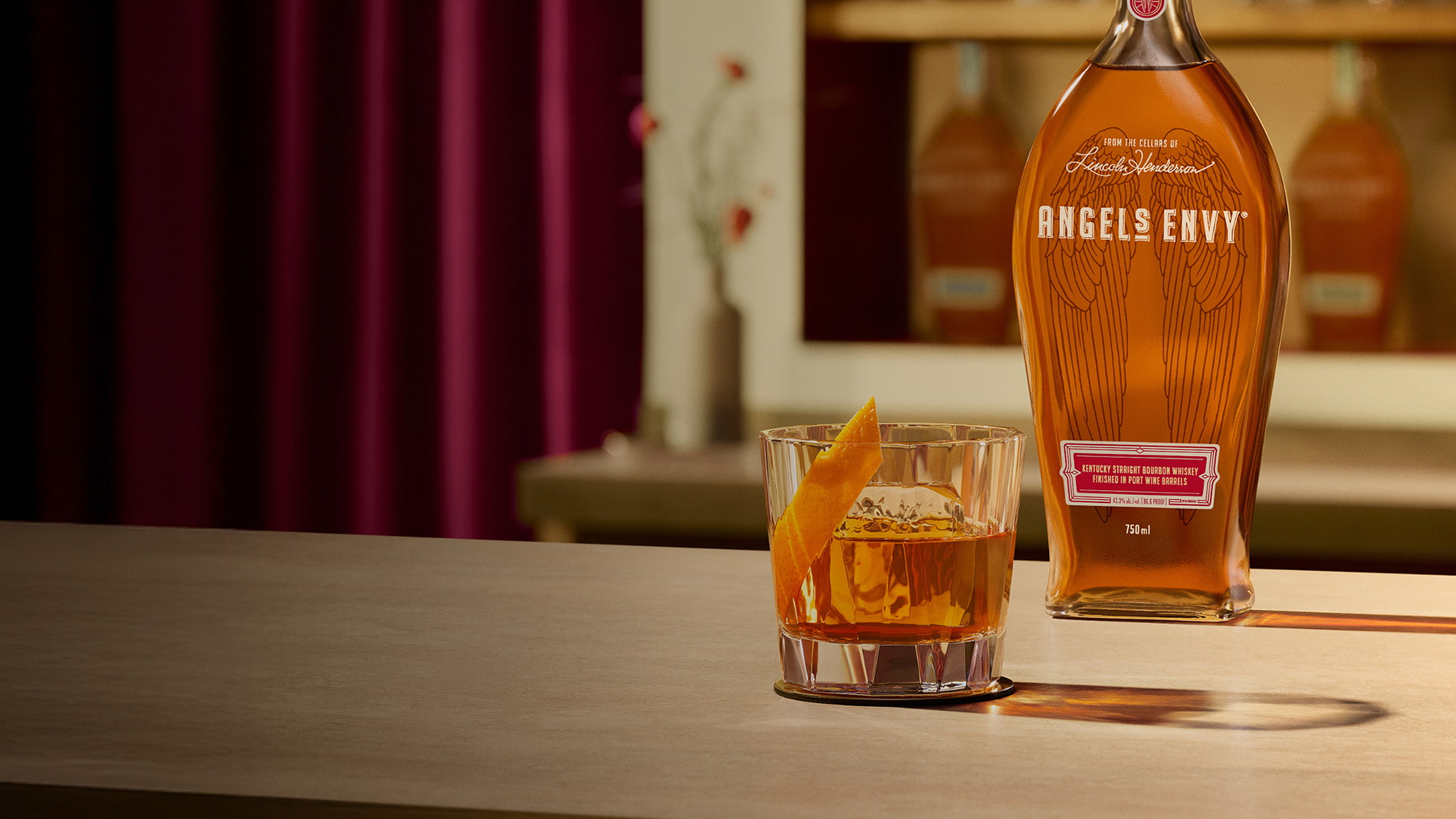 On a light wooden table in a modern bar setting with rich ruby design detailing, an old fashioned cocktail with an orange garnish is presented beside a bottle of Angel's Envy Bourbon.