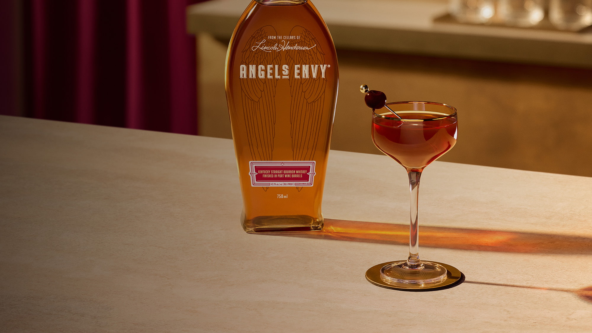 On a light wooden table in a modern bar setting with rich ruby design detailing, a Manhattan cocktail with an cherry garnish is presented beside a bottle of Angel's Envy Bourbon.