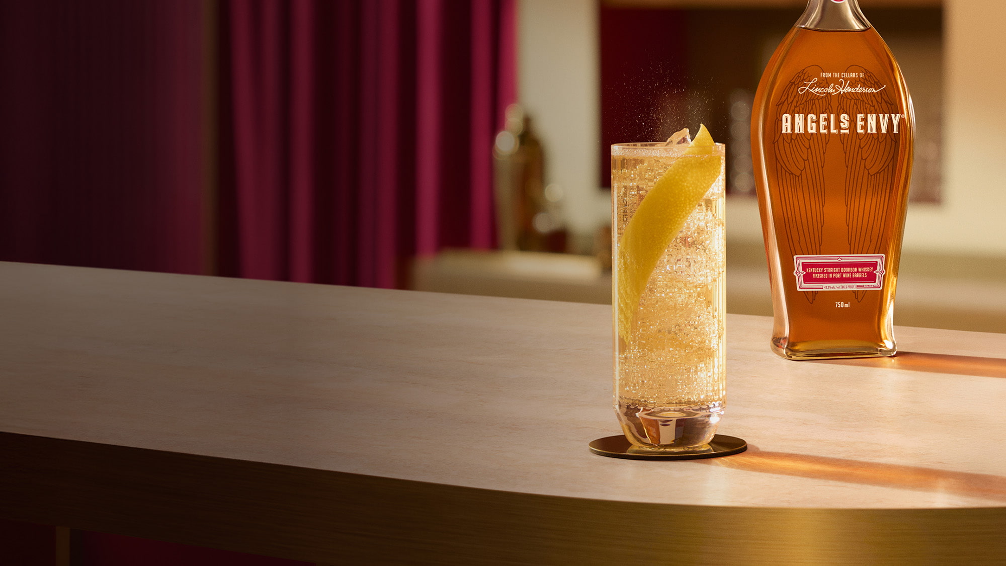 On a light wooden table in a modern bar setting with rich ruby design detailing, a highball cocktail with a lemon peel garnish is presented beside a bottle of Angel's Envy Bourbon.