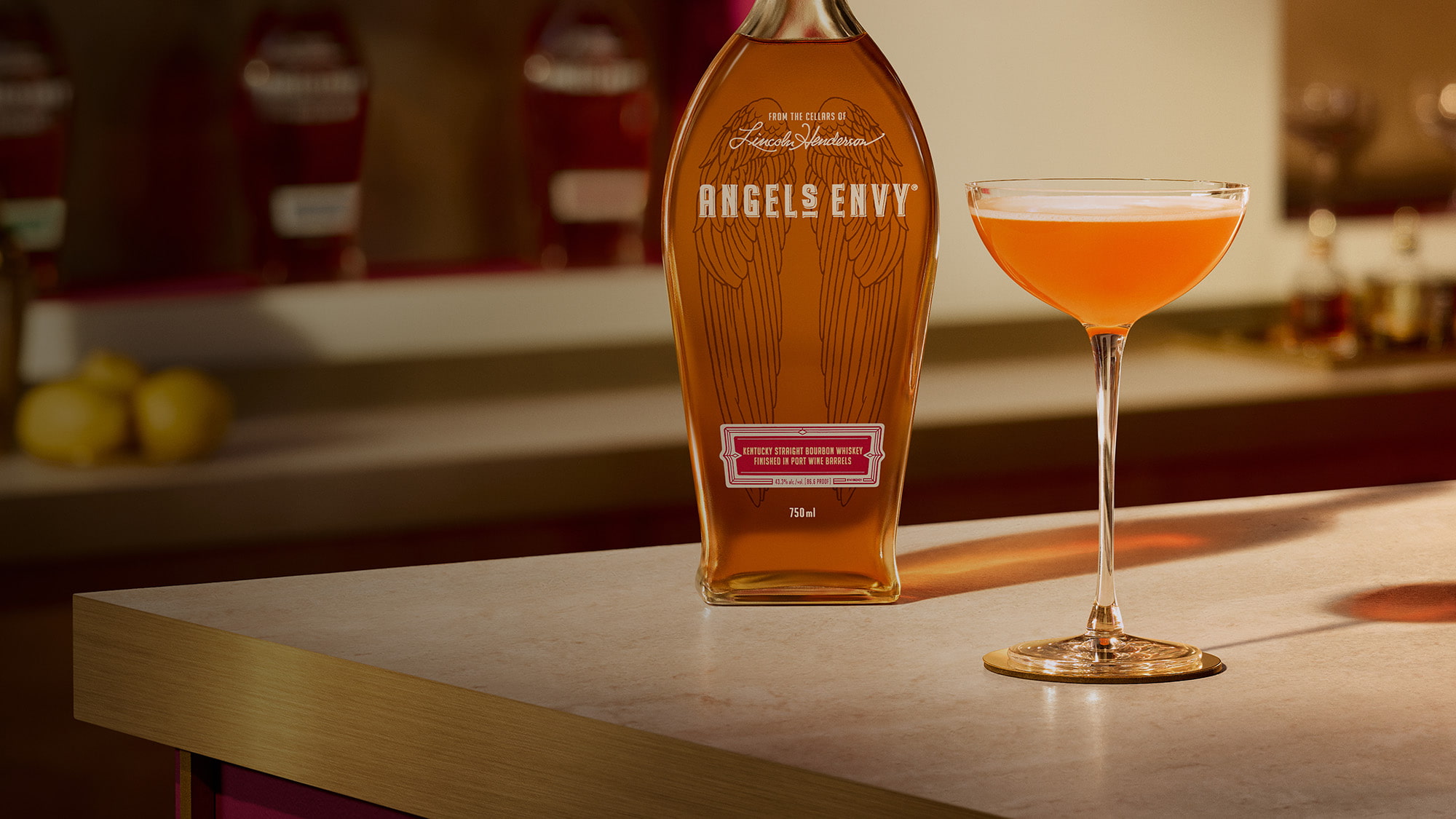 On a light wooden table in a modern bar setting with rich ruby design detailing, a paper plane cocktail is presented beside a bottle of Angel's Envy Bourbon.