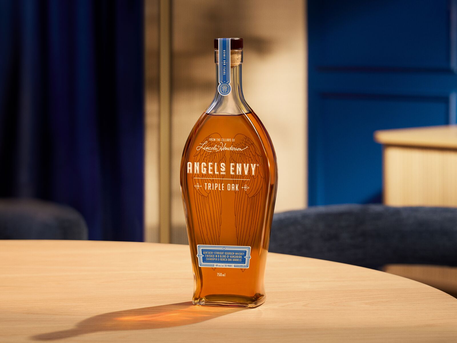 A 750 mL bottle of Angel's Envy Triple Oak with a signature wings graphic in black is presented in a modern bar setting with blue and bright wood design detailing.