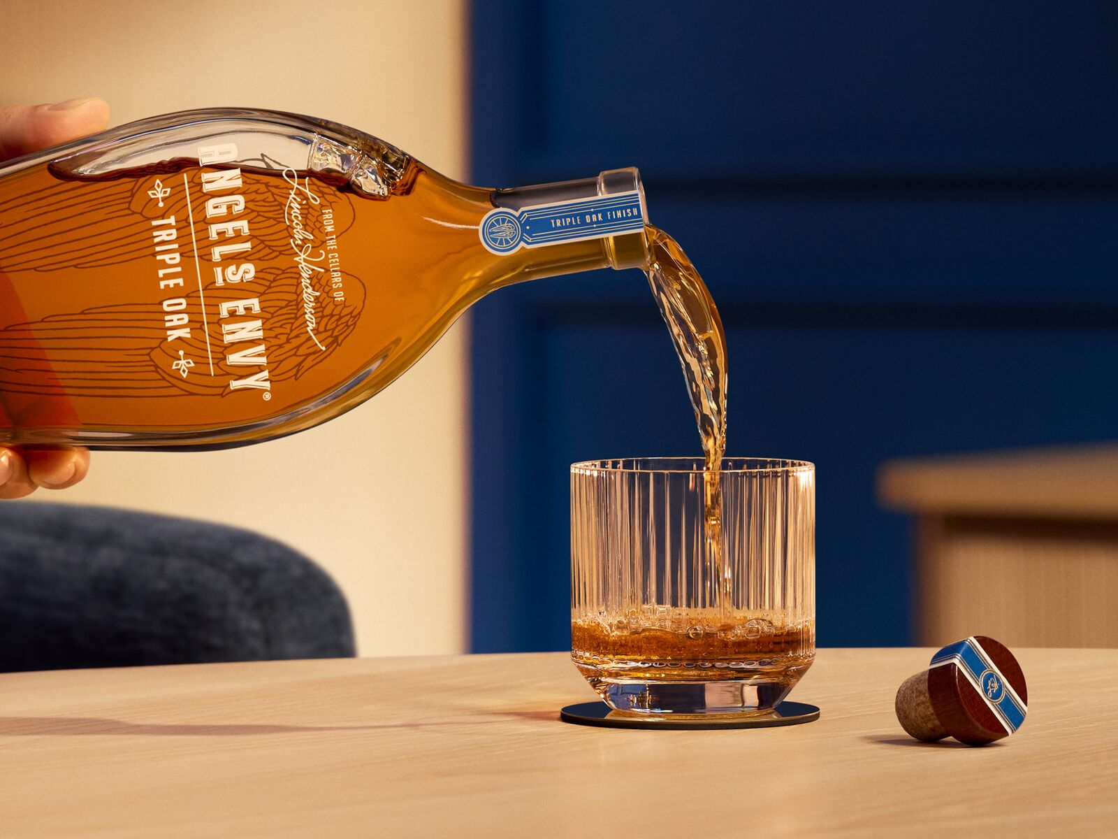 A 750 mL bottle of Angel's Envy Triple Oak with a signature wings graphic in black is poured into a rocks glass, set in a modern bar with blue and bright wood design detailing.