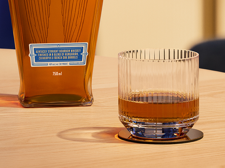 A neat glass of Angel's Envy Triple Oak bourbon with the bottle resting in the background on a white textured table.