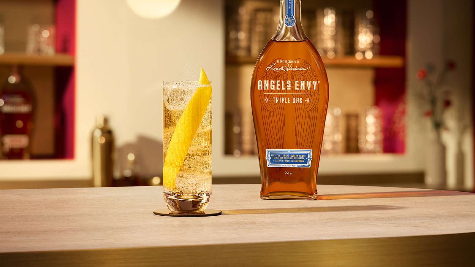 On a white, textured table-top in a modern bar setting with rich ruby design detailing, a highball cocktail with a lemon peel garnish is presented beside a bottle of Angel's Envy Triple Oak Bourbon.