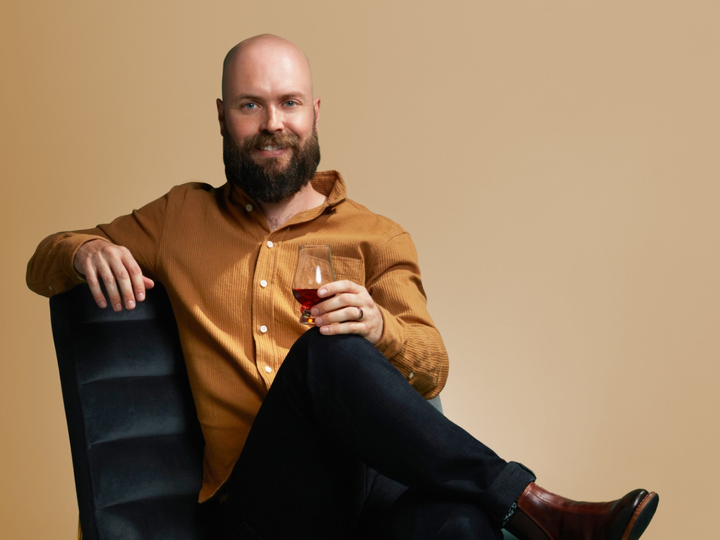 A smiling, bearded Master Distiller Owen Martin sitting cross-legged with a near glass of The Angel’s Envy 2024 Cask Strength Kentucky Straight Bourbon Whiskey.