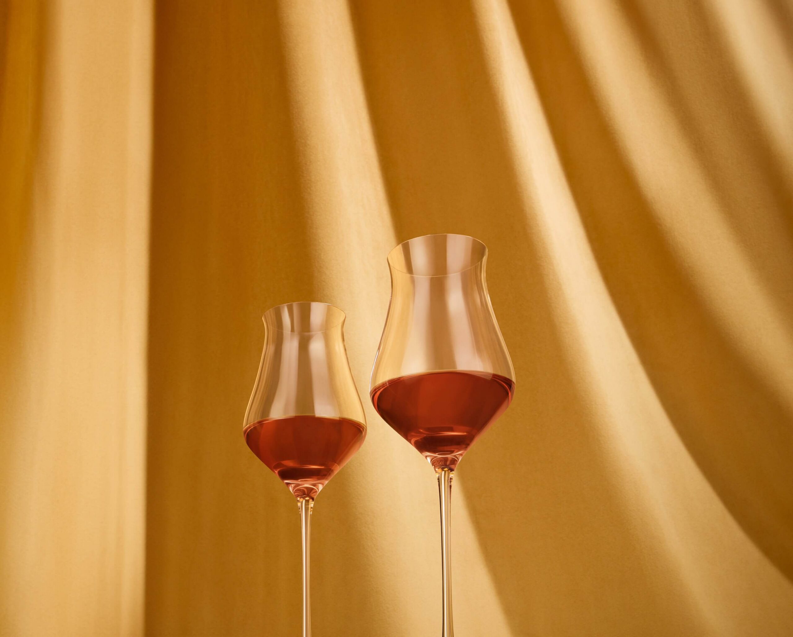 Shot from below, two towering long-stemmed tasting glasses are presented in front of a golden textured curtain.