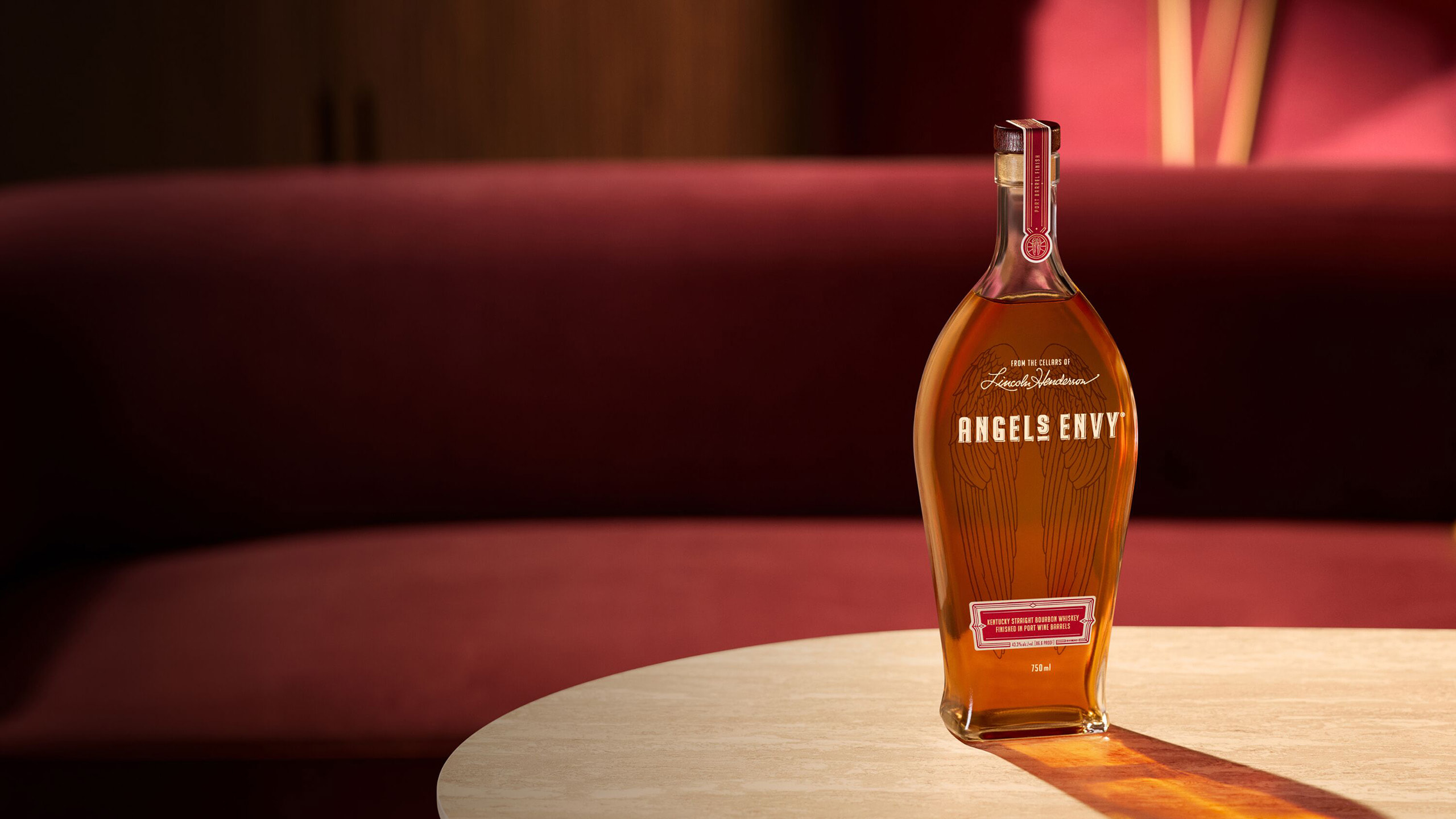 A bottle of Angel's Envy Kentucky Straight Bourbon Whiskey Finished in Port Wine Barrels on a white textured table in a room with deep ruby decor.