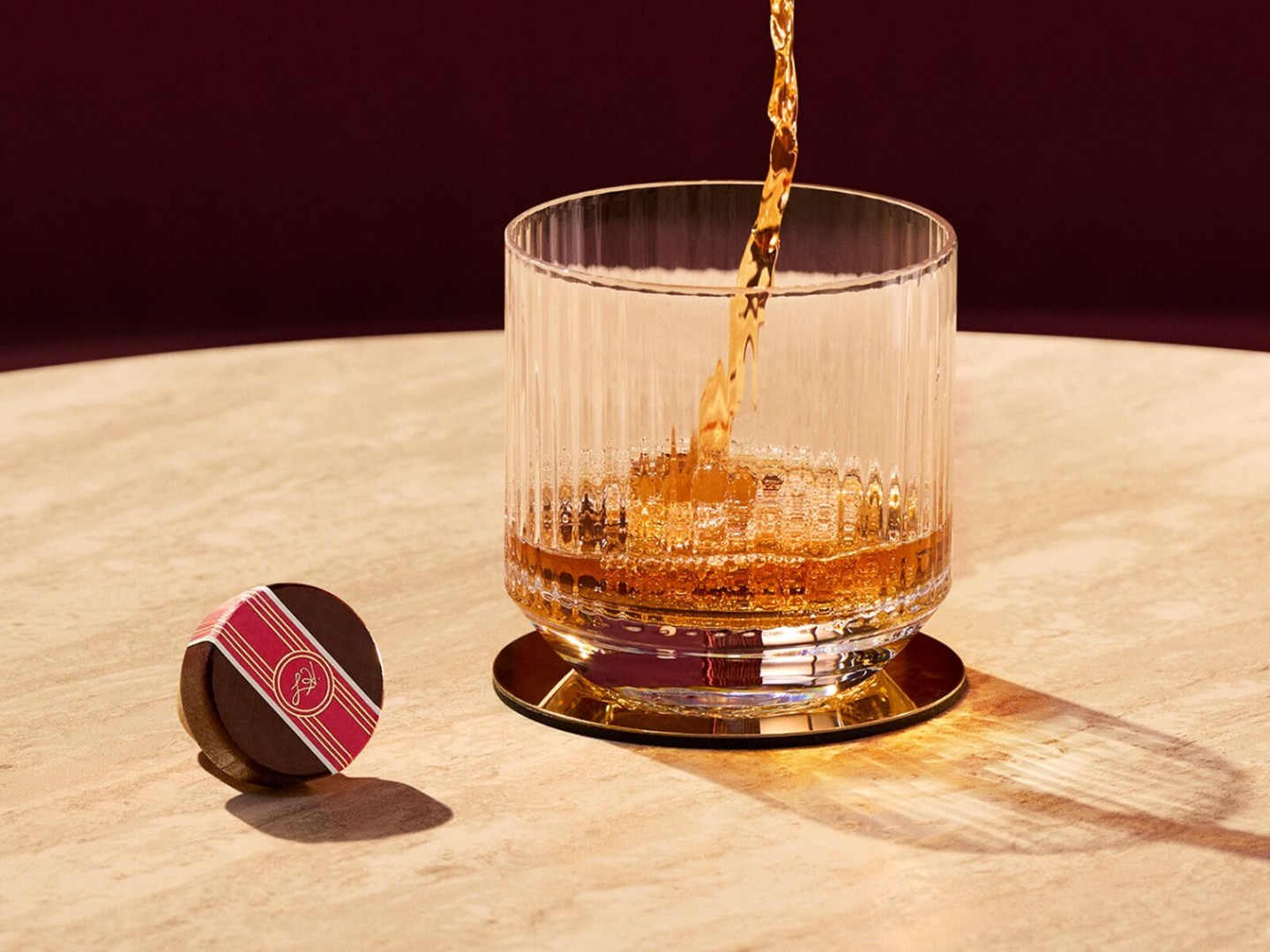 A rocks glass receives a pour of Angel's Envy Bourbon on a white, textured table-top with a cork resting to the side.