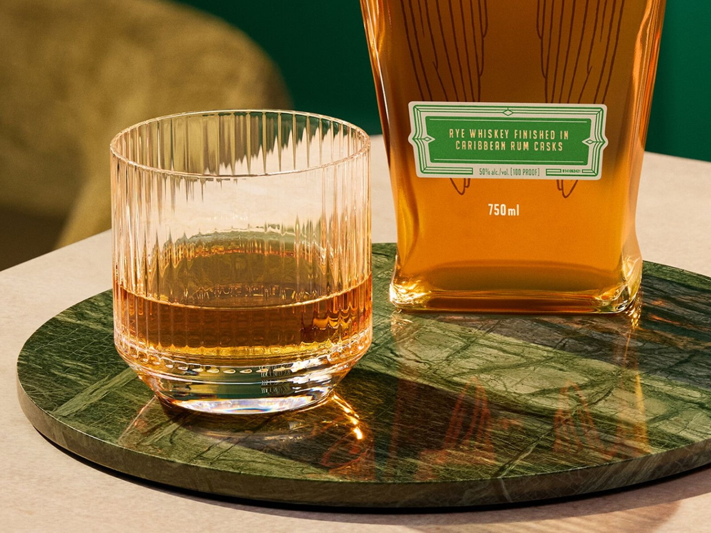 The Angel's Envy Rye Finished in Caribbean Rum Casks bottle set on a metallic green plate, with a neat glass of rye to the side.