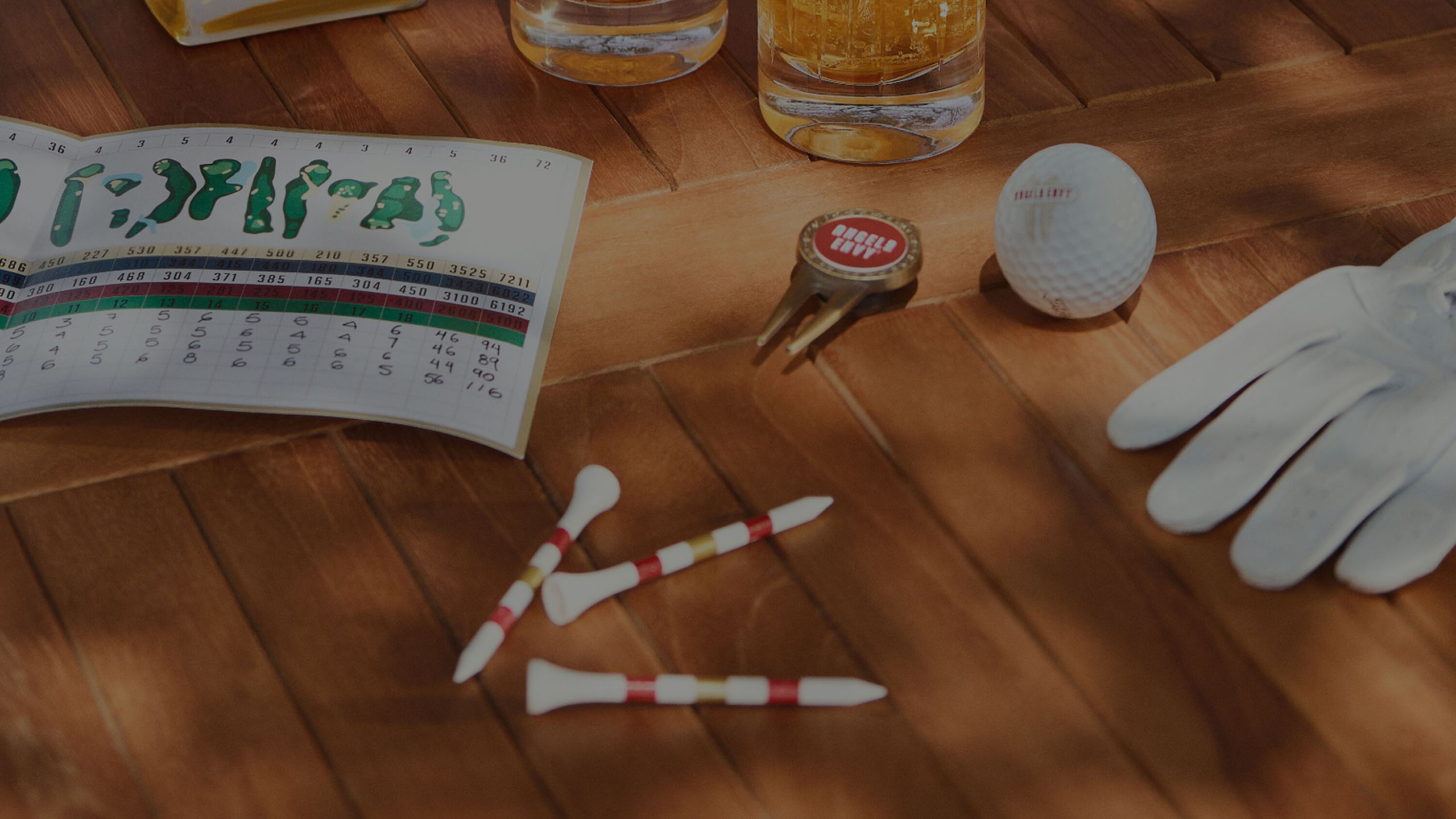The text Whiskey Gifts for Golf Lovers overlays a wooden outdoor table at the course feauring Angel’s Envy golf tees, a ball marker, a branded golf ball, a score card and cocktails flicker with sunlight.