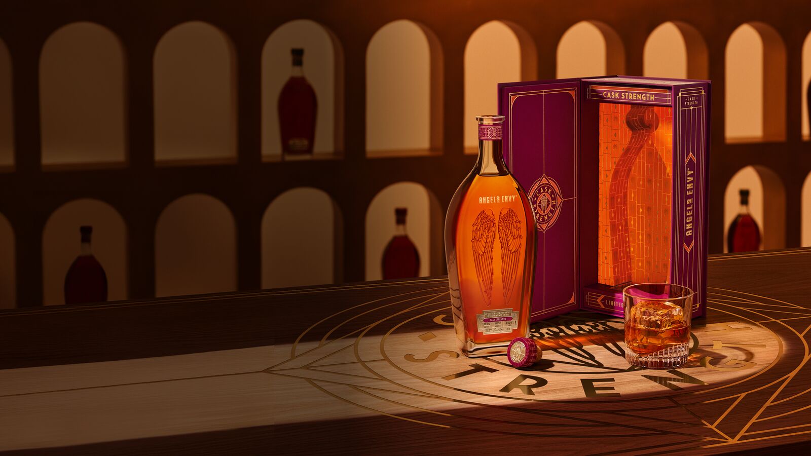 Angel’s Envy 2026 Cask Strength Bourbon bottle, the limited-edition presentation box in ruby and gold, and a rocks glass of whiskey presented together on a logo-engraved table in the Cask Archives room.