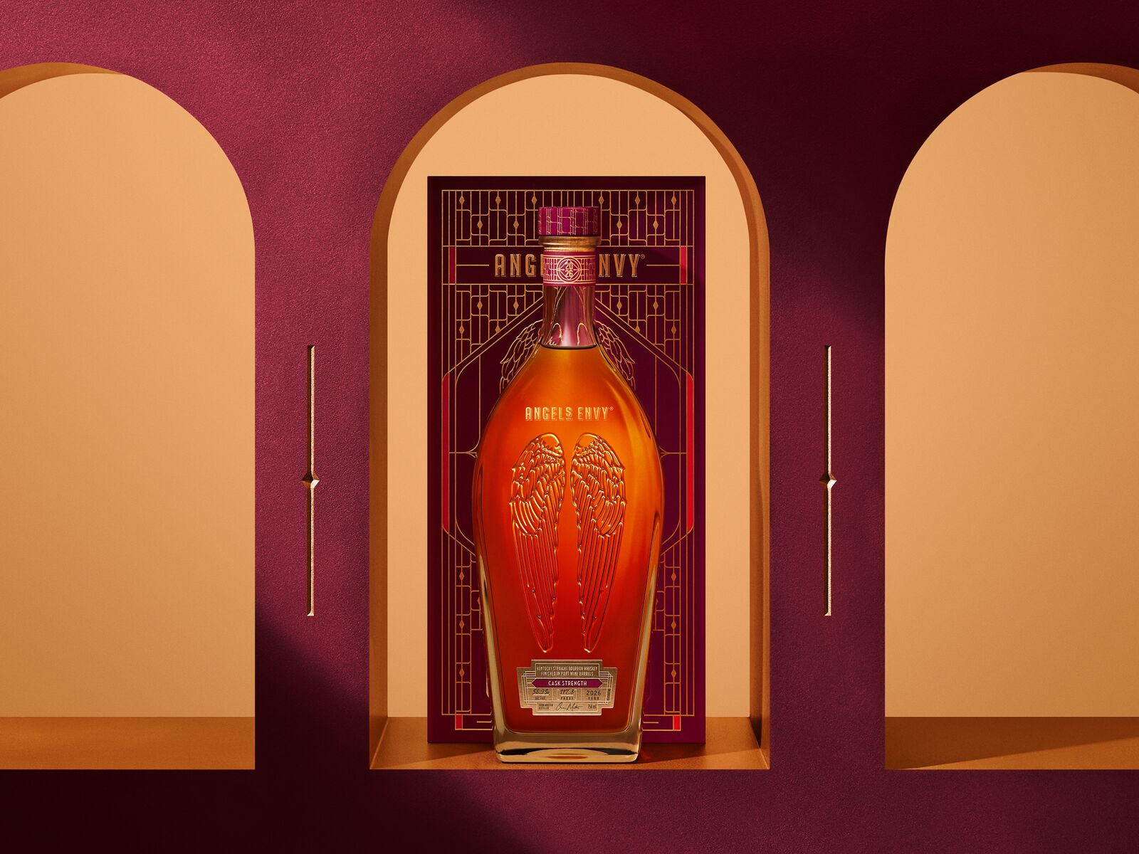 Angel’s Envy 2026 Cask Strength Bourbon bottle displayed in front of the limited-edition presentation box, framed by an arched niche in the Cask Archives room with a ruby background and golden archway.