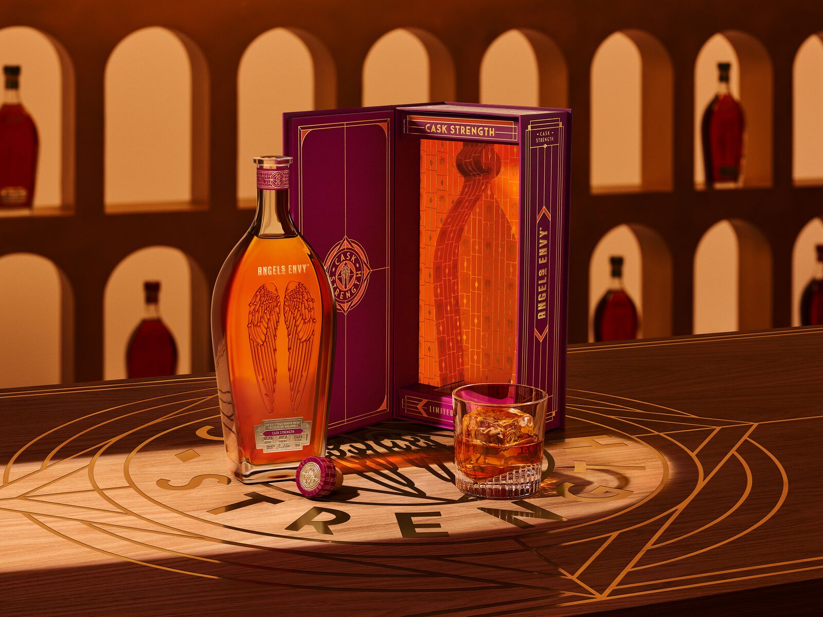 Angel’s Envy 2026 Cask Strength Bourbon bottle, the limited-edition presentation box in ruby and gold, and a glass of whiskey displayed together on a logo-engraved table in the Cask Archives room.