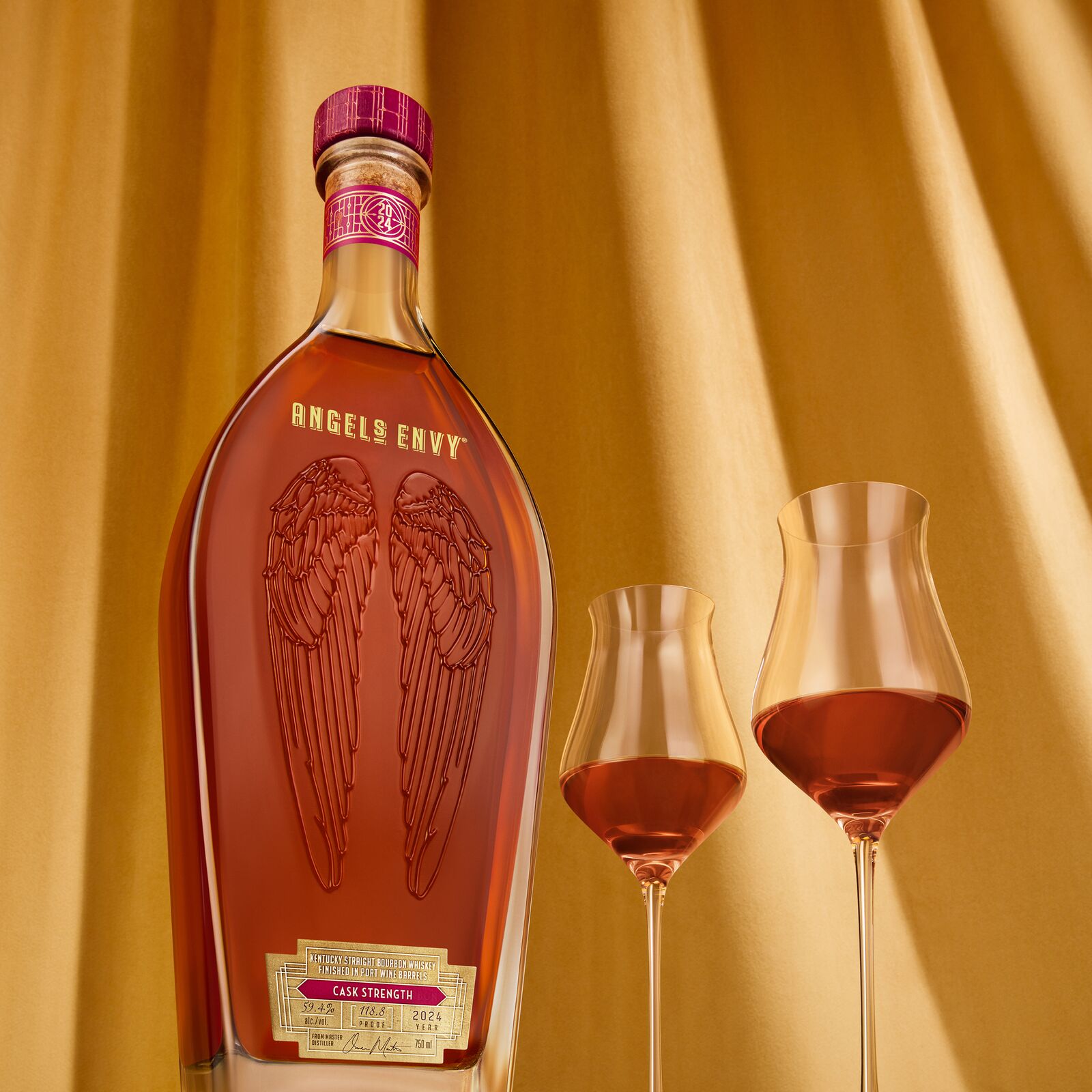 Shot from below, a towering bottle of Angel’s Envy Cask Strength Bourbon 13th Edition with two long-stemmed tasting glasses are presented in front of a golden textured curtain.