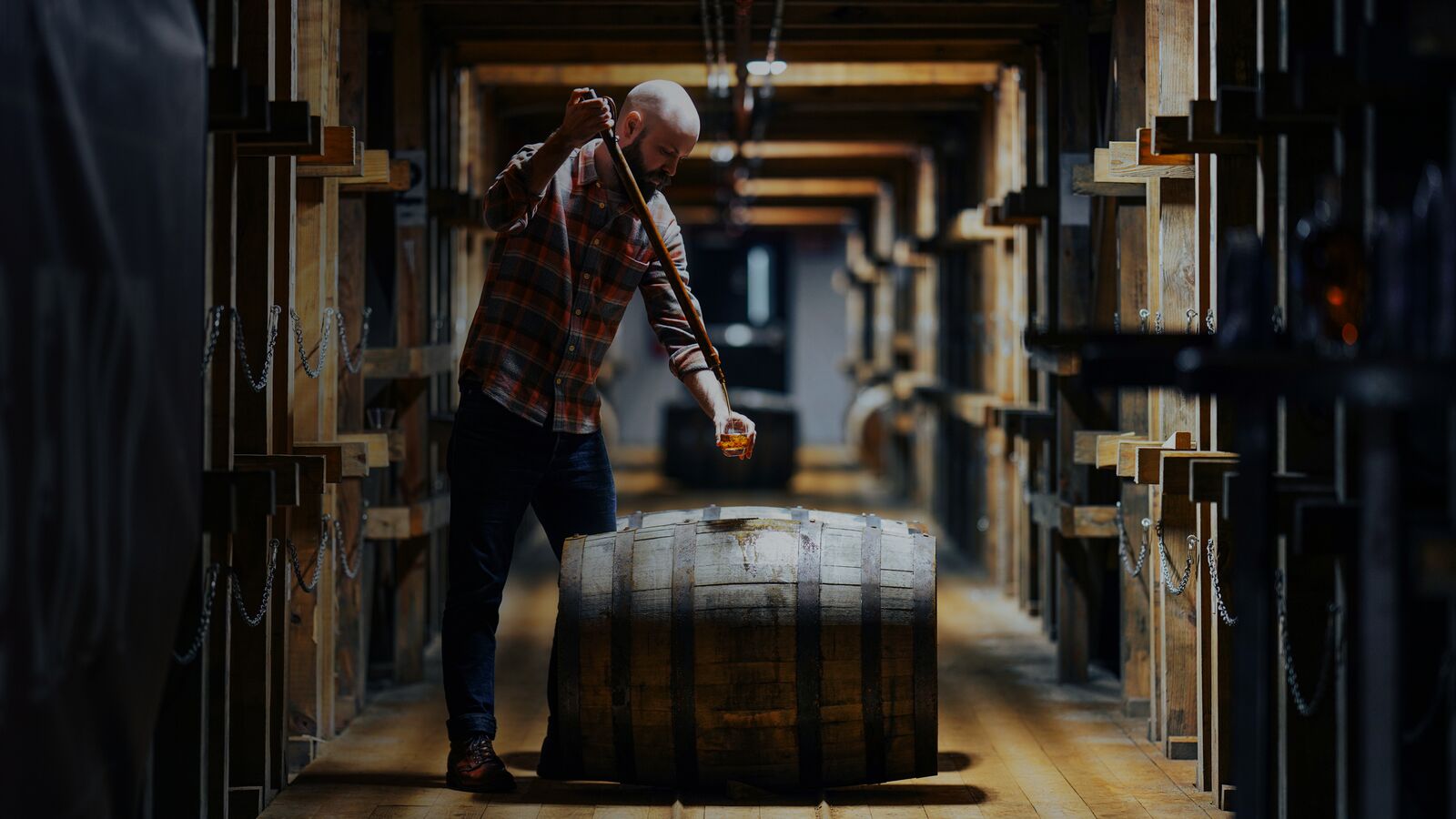 A spotlit Master Distiller Owen Martin barrel thieving in the Angel’s Envy rickhouse between endless barrel shelves.