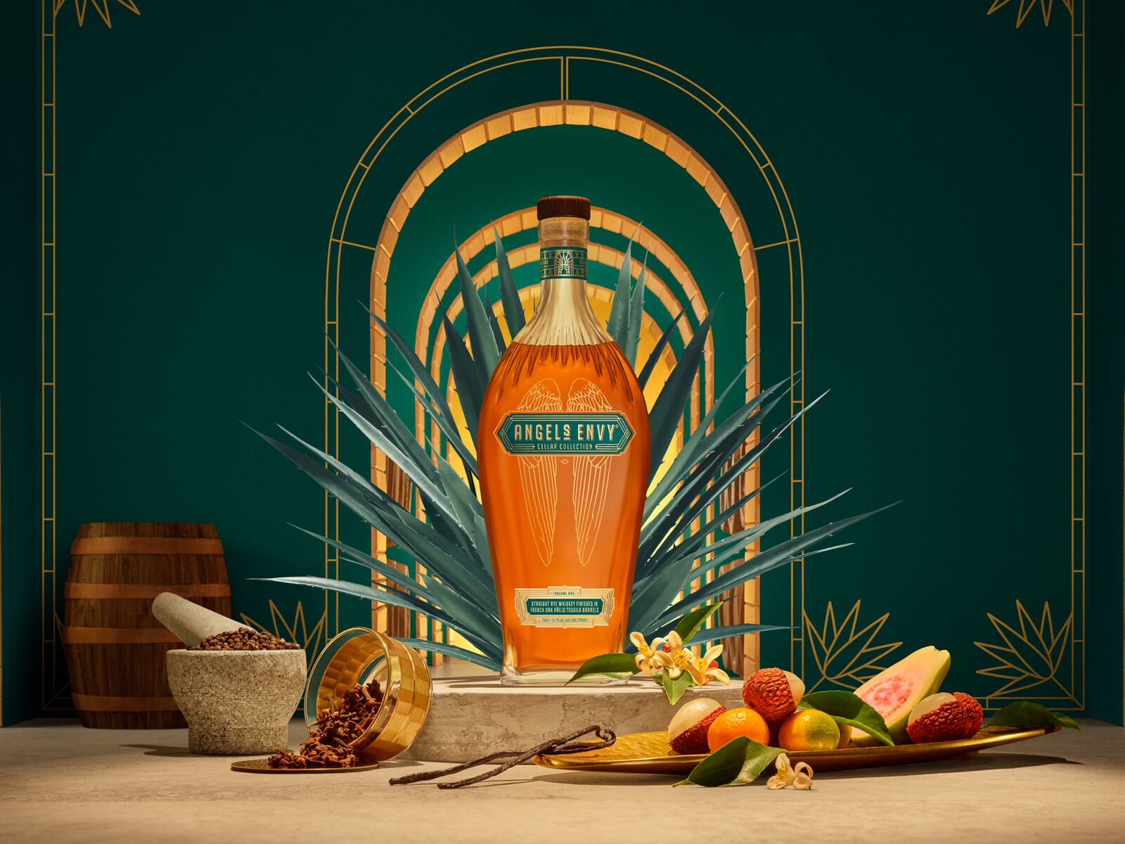 A spotlit Angel’s Envy Cellar Collection Vol. 5 bottle displayed with its emerald green and gold presentation box, set on a textured stone pedestal against a green and gold architectural backdrop featuring an agave plant, spices, and fruit.