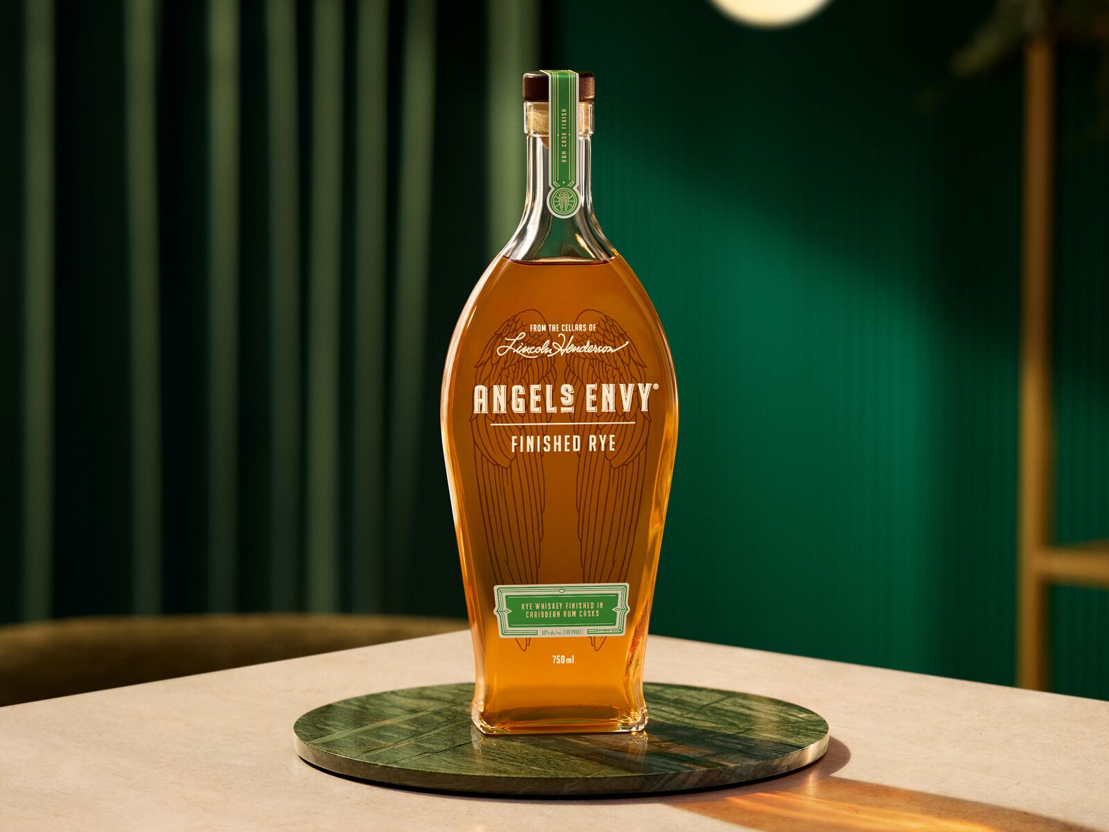A spotlit bottle of Angel’s Envy Rye set on a metallic green plate, presented within within a modern room featuring deep green walls and velvet curtains.