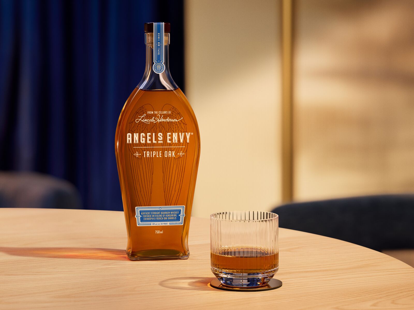 Set inside warmly lit contemporary space, a bottle of Angel’s Envy Triple Oak bourbon and a neat glass of its liquid sits atop a light wooden table.
