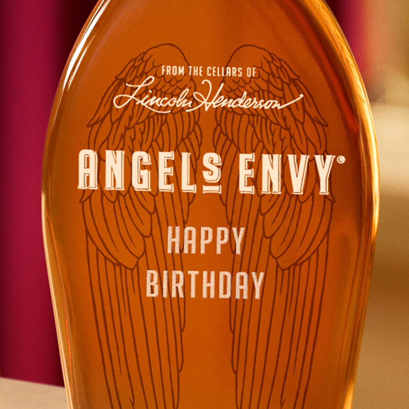 A warmly lit bottle of Angel’s Envy Bourbon with this signature wings in black, featuring a custom engraving that reads “Happy Birthday.”