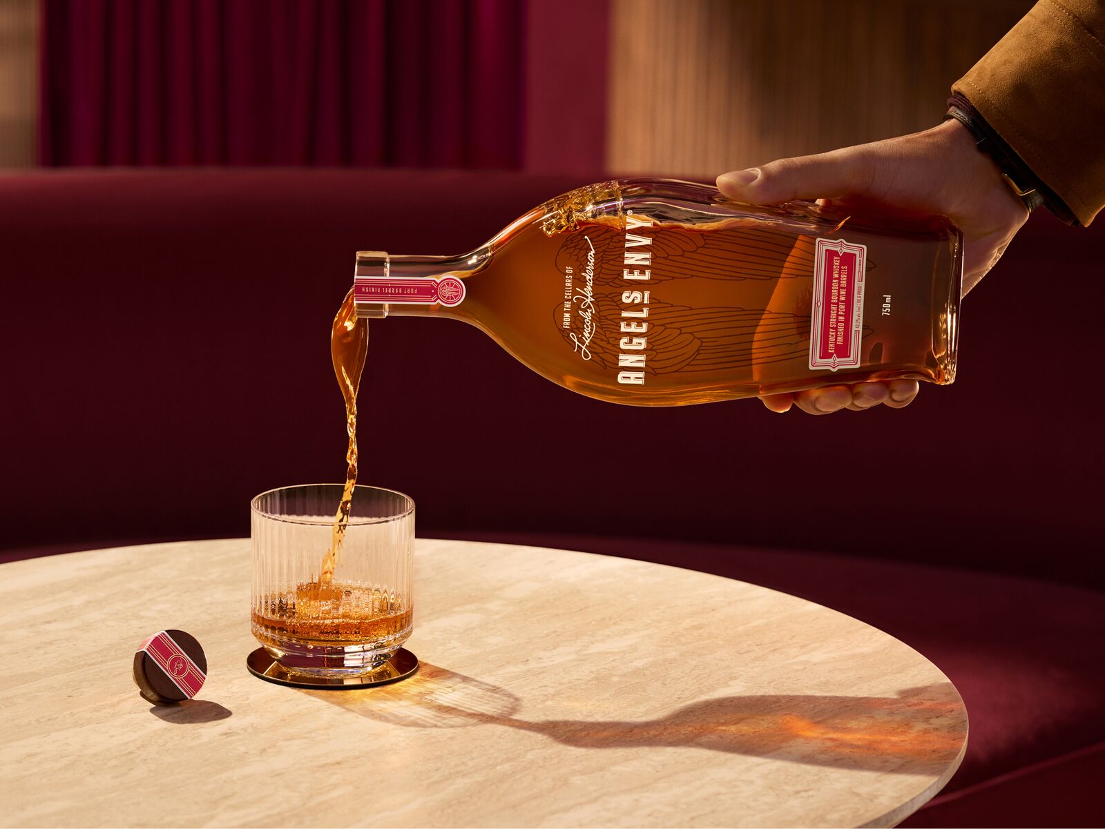 A man’s hand pouring into a rocks glass with a bottle of Angel’s Envy Signature Series bourbon with the labeled cork at its side, set on top of a warmly lit modern table.