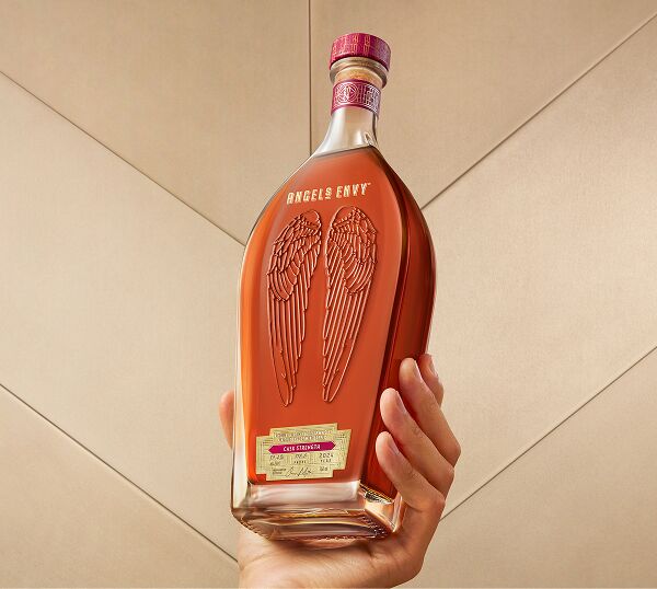A hand presenting a bottle of Angel’s Envy Cask Strength Bourbon 13th Edition in front of a tiled light beige wall.