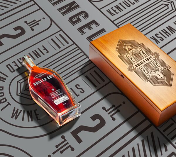 A laid-down bottle of the Angel’s Envy 2022 Cask Strength Bourbon Whiskey Finished in Port Wine Barrels and its logo-engraved presentation box in a light brown wood, set on a logo-printed background in black and gray.