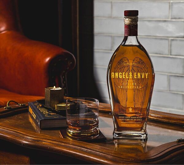 The Cellar Collection Vol. 2 bottle on a polished wooden drink tray featuring a rocks glass, a black leather-bound book, and a candle with a black vessel; resting in front of a rustic ruby leather armchair.
