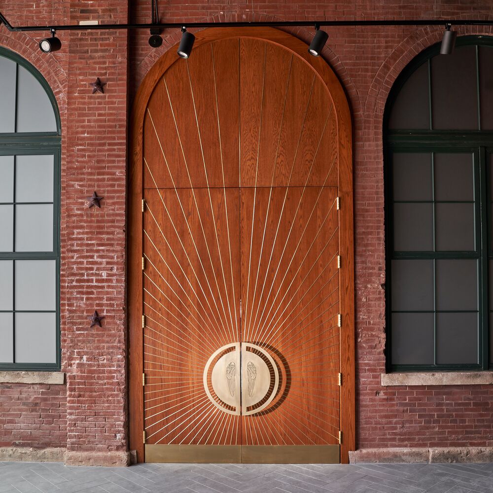 A central view of the grand wooden, arched doors featuring a golden, wing-engraved hardware and surrounding red brick walls within the Angel’s Envy Distillery in Louisville, Kentucky.