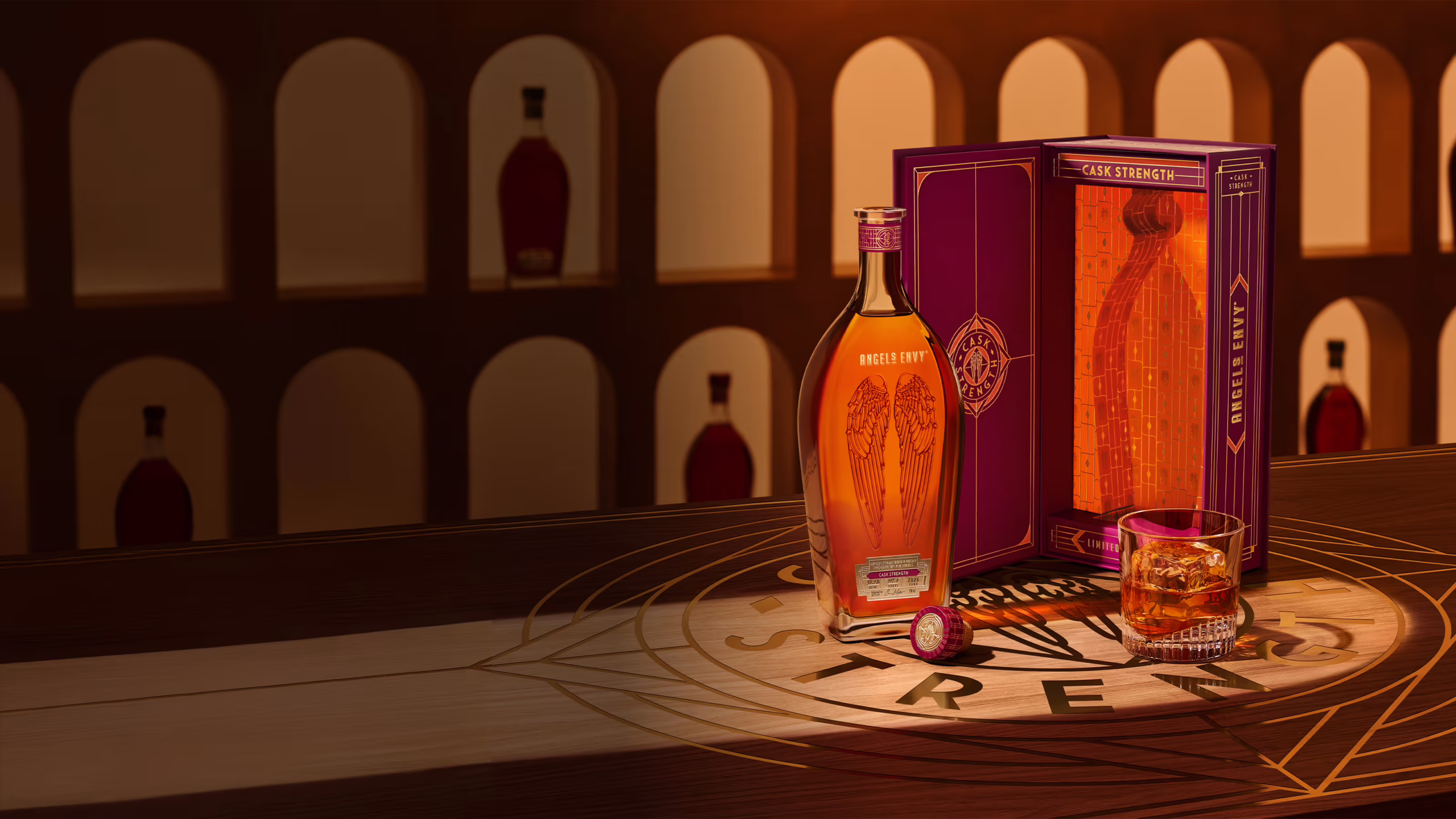 Angel’s Envy 2026 Cask Strength Bourbon bottle, the limited-edition presentation box in ruby and gold, and a rocks glass of whiskey presented together on a logo-engraved table in the Cask Archives room.