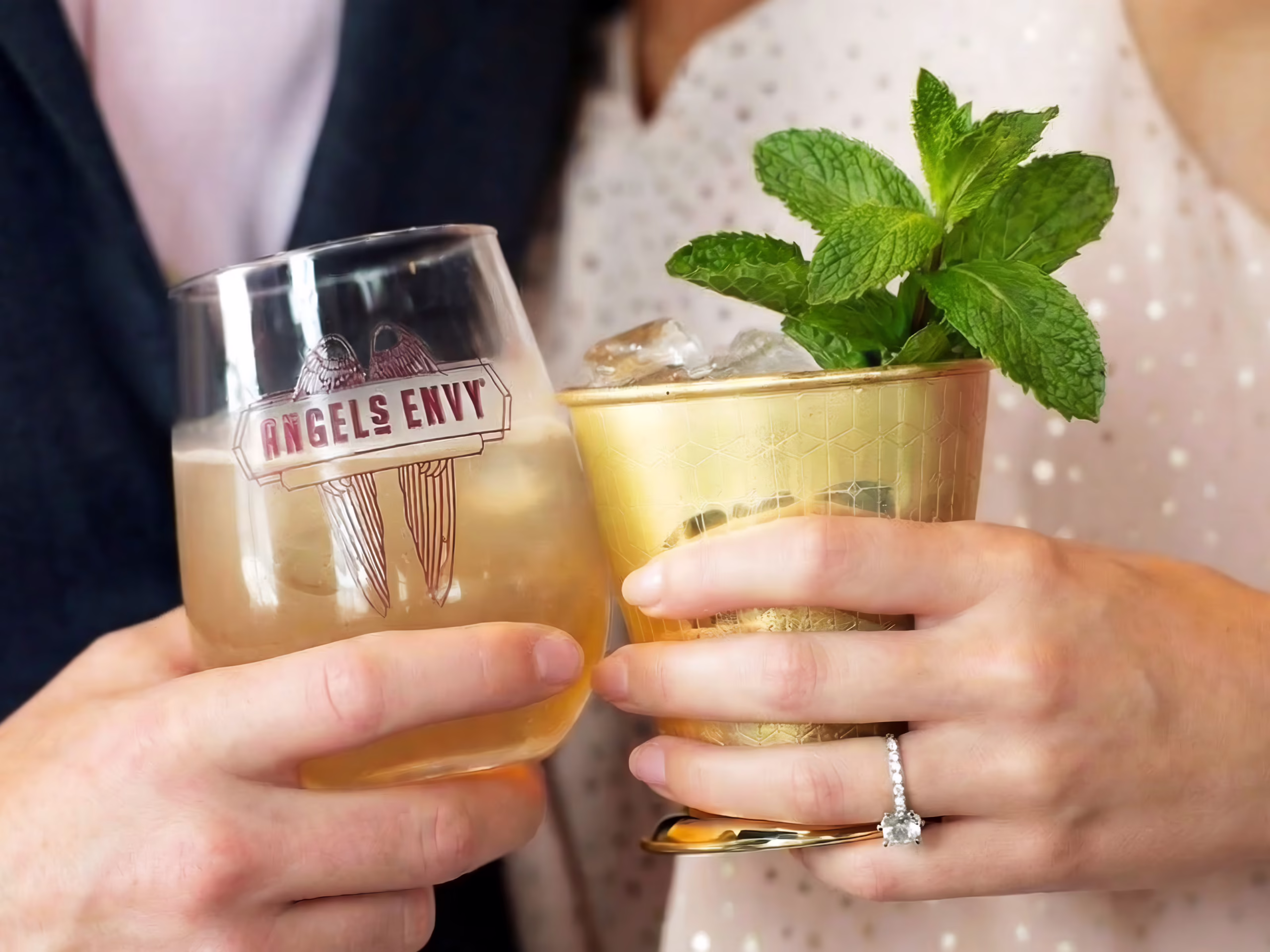 A close-up of a man and woman’s hands toasting their cocktails at the Angel’s Envy Distillery, featuring a whiskey sour in an Angel’s Envy brand glass and frosted Mint Julep.