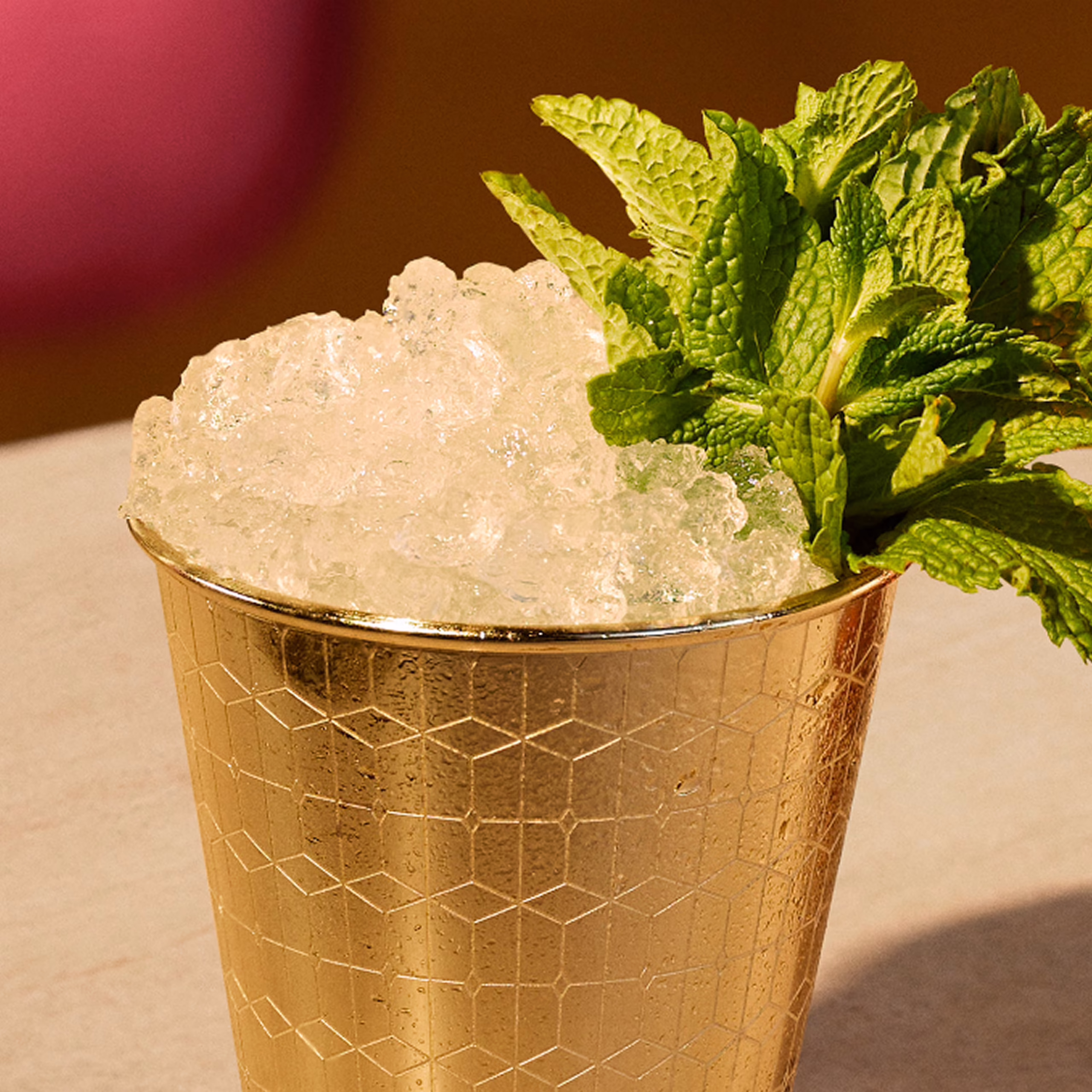 Set in a warmly-lit modern bar with ruby chairs and white tabletop, an iced Mint Julep cocktail is presented in a golden signature Angel’s Envy Mint Julep cup.