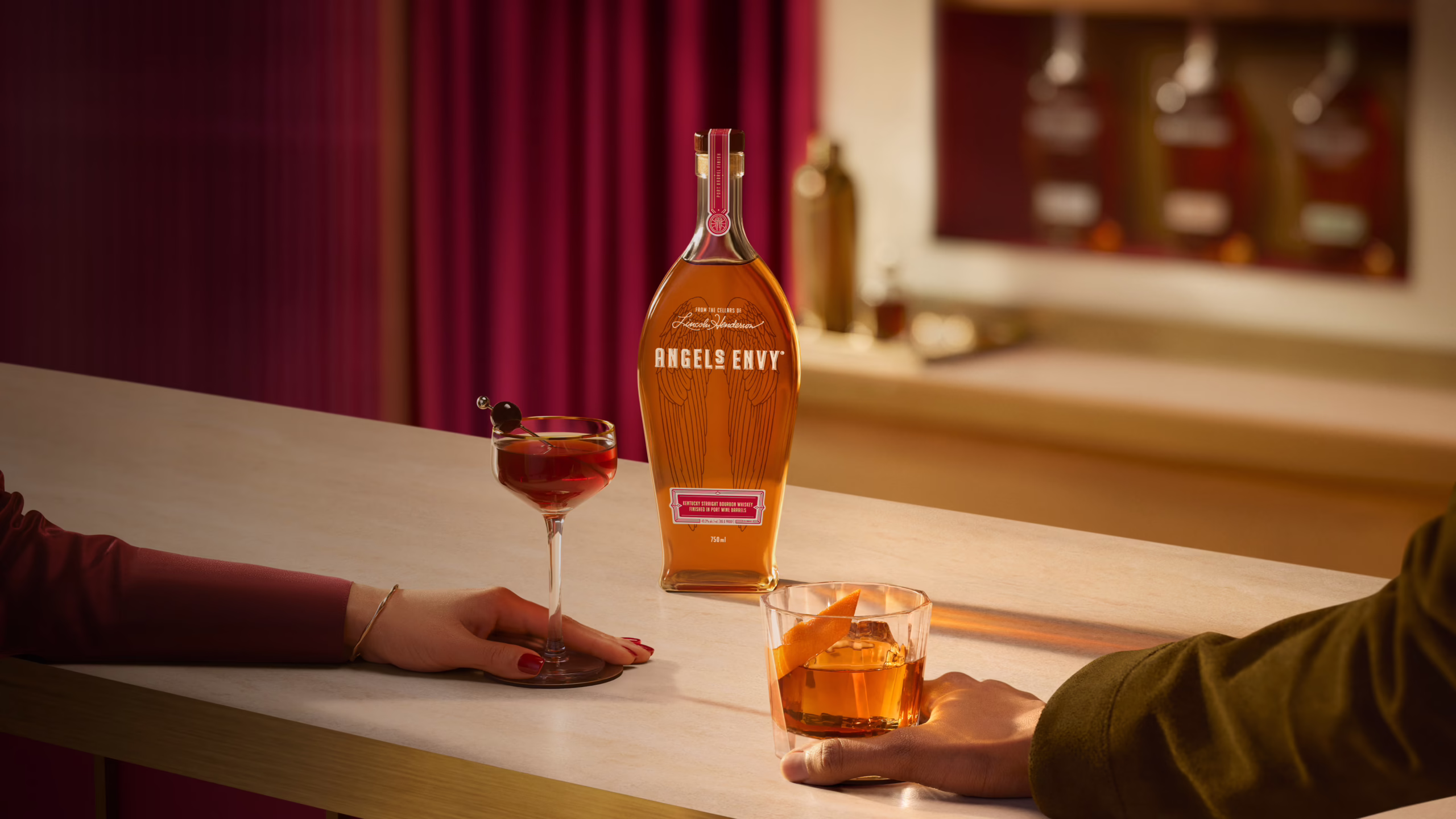 Set on a white marble bar in the Angel’s Envy Distillery, a couple enjoys a Manhattan and an Old Fashioned cocktail with a bottle of Angel’s Envy bourbon on display.