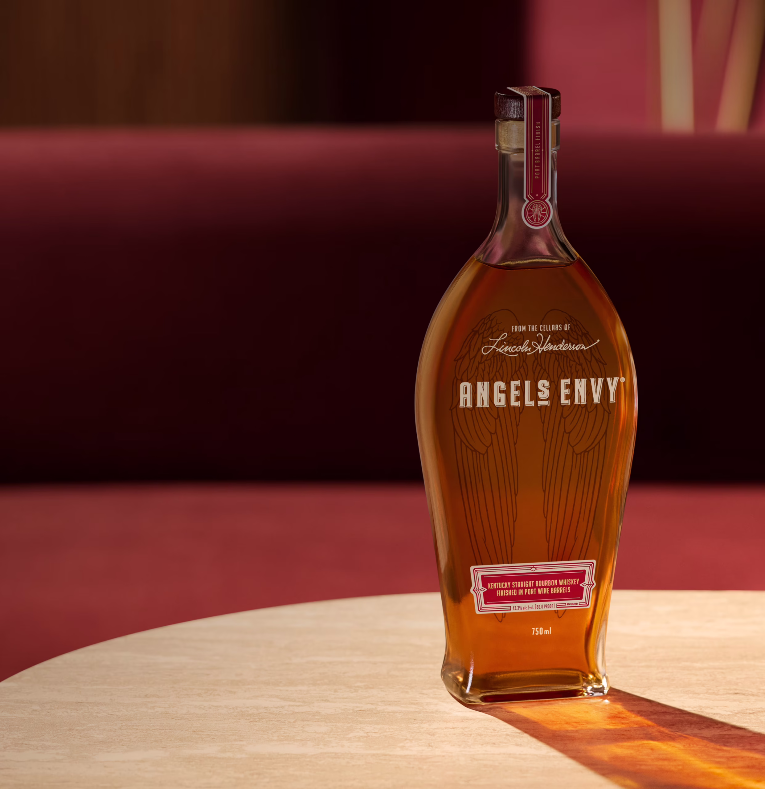 Set in a warmly-lit modern bar with ruby decor and white tabletop, a bottle of Angel’s Envy 2026 Cask Strength Bourbon is presented and casts a golden shadow.