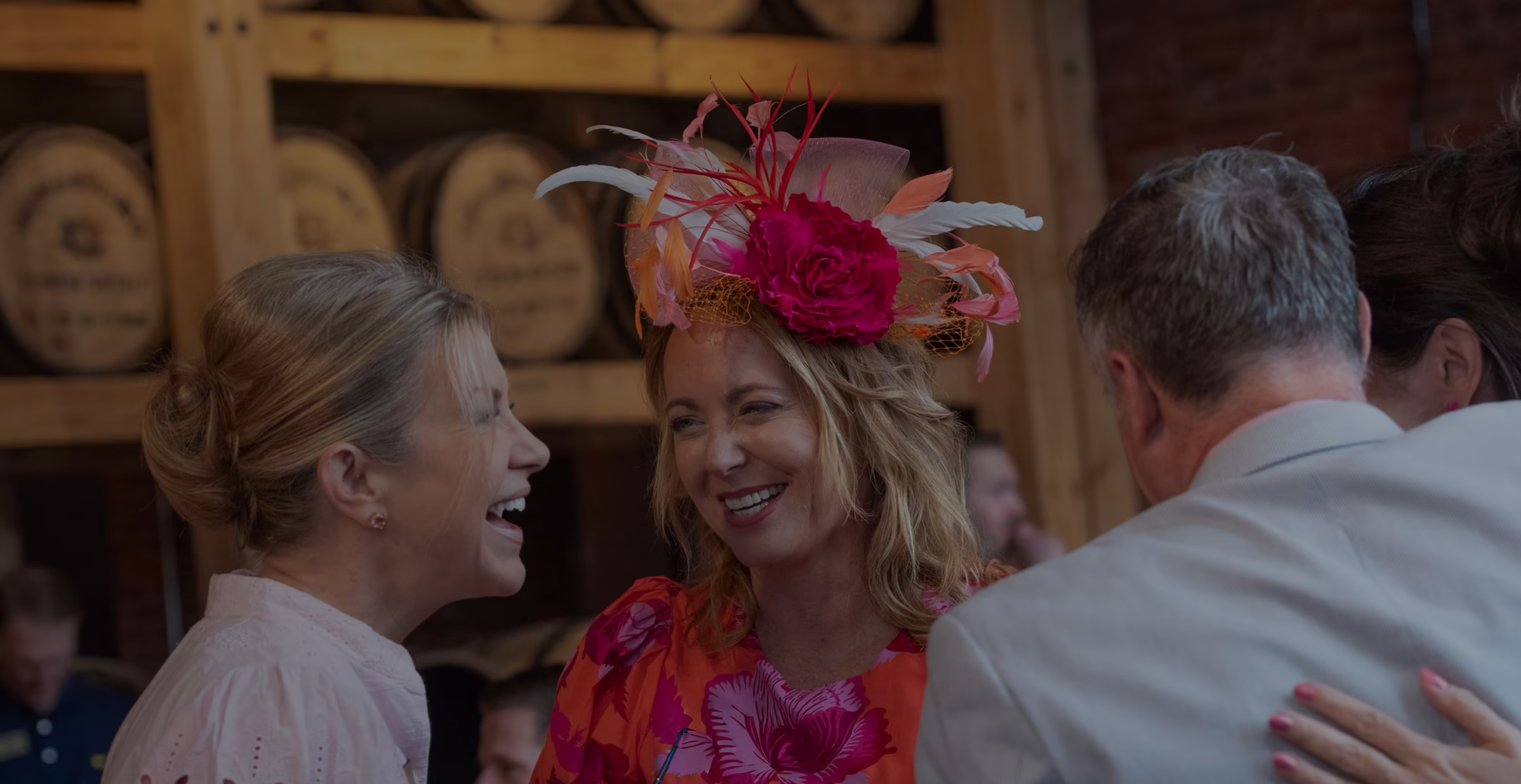 At the Angel’s Envy Distillery, a group of joyful guests celebrate the occasion in classic Derby attire, featuring feather-adorned fascinators and floral designs.