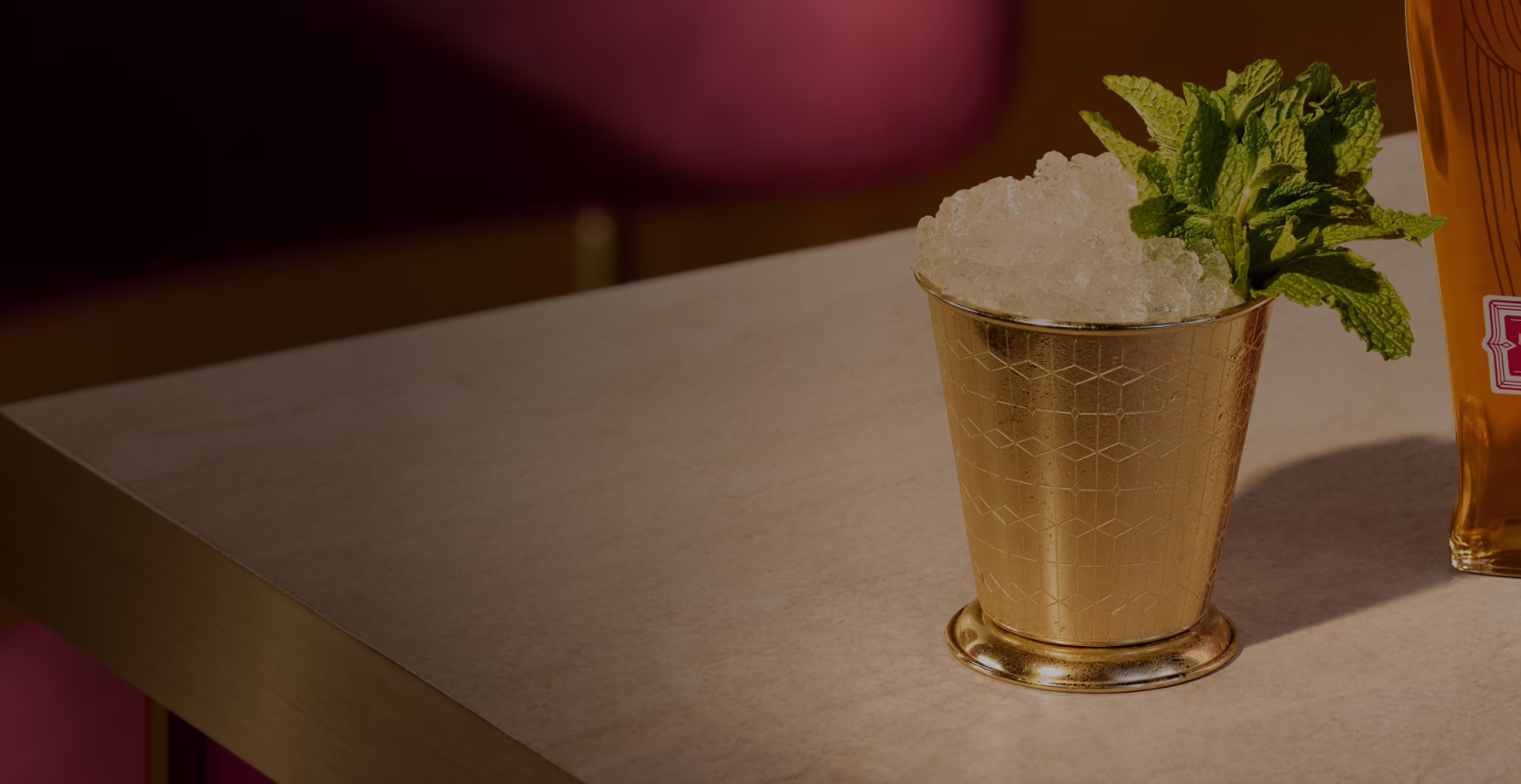 Set in a warmly-lit modern bar with ruby chairs and white tabletop, an iced Mint Julep cocktail is presented in a golden signature Angel’s Envy Mint Julep cup.