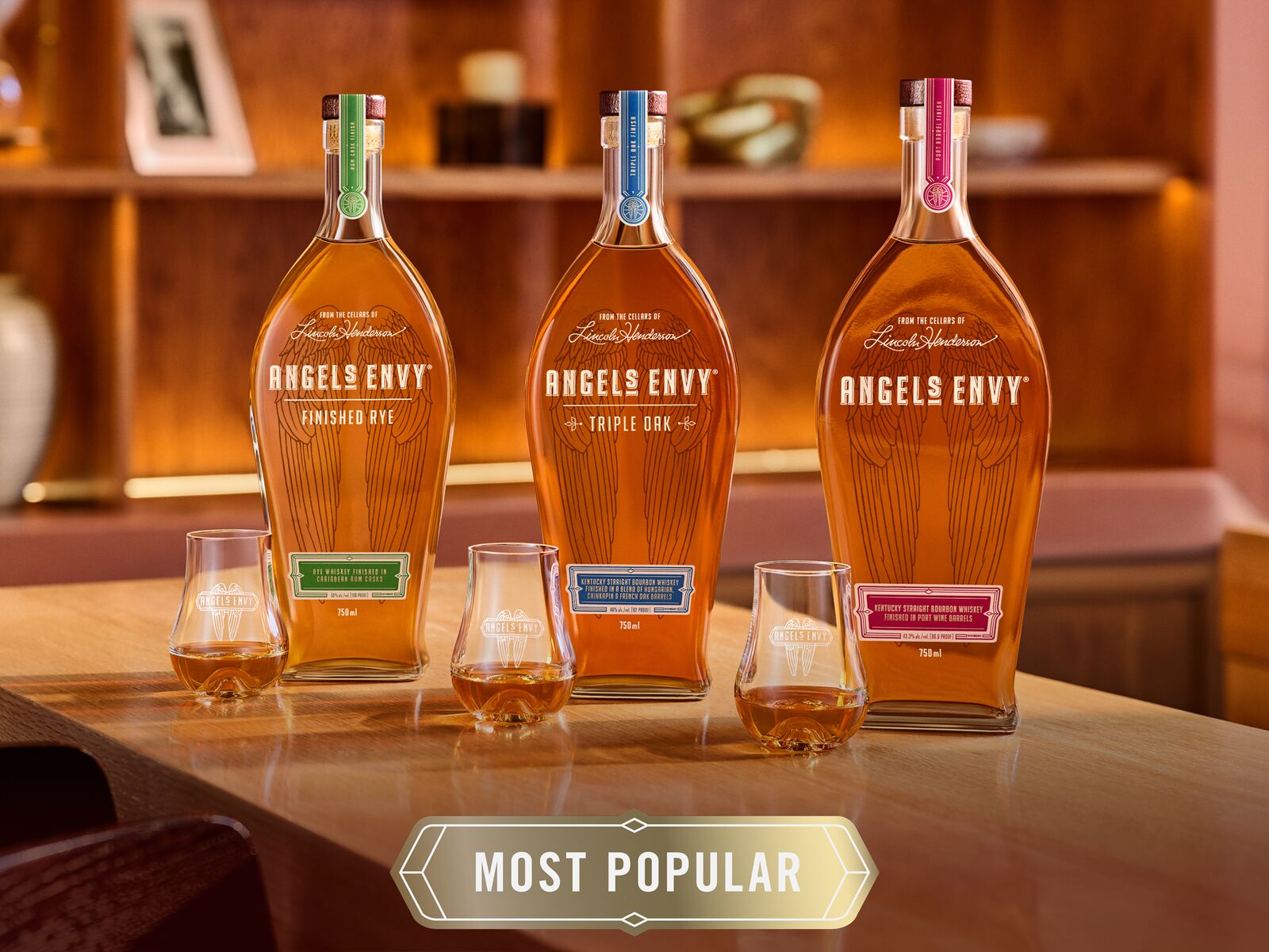 Set in the modern, wood-detailed tasting room at the Angel's Envy Distillery, the three Angel's Envy Signature Series whiskey bottles are lined up beside three tasting glasses with the words "Most Popular" in a golden art deco graphic.