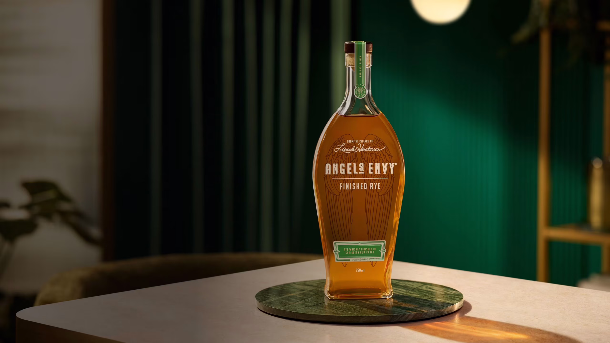 Presented within a modern room featuring deep emerald walls and velvet curtains, a bottle of Angel's Envy Rye Finished in Caribbean Rum Casks rests on a metallic green plate.