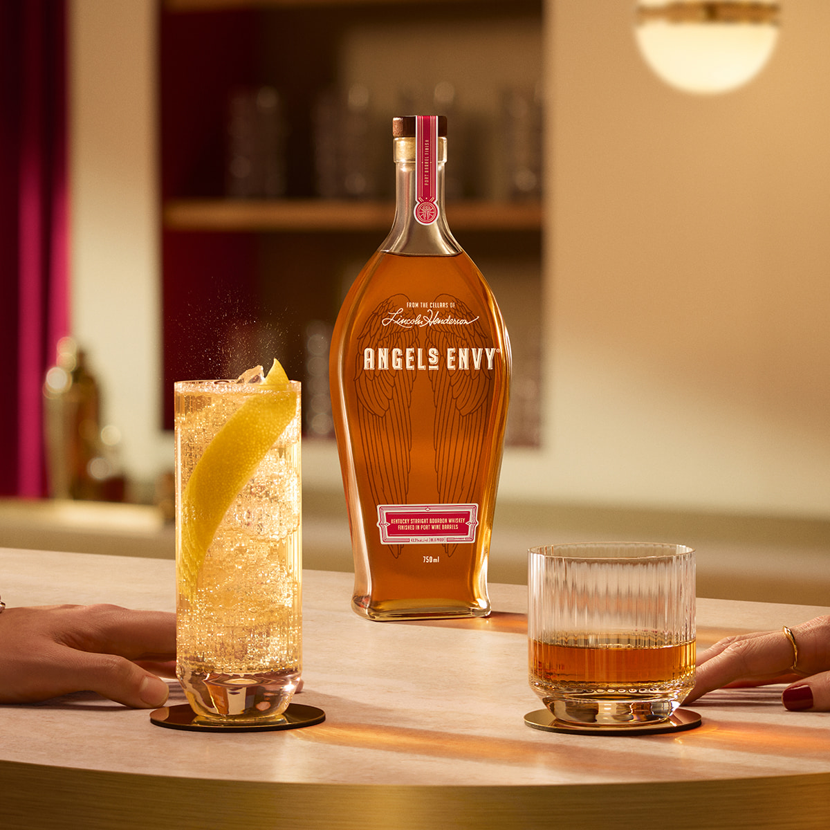 On a light wooden table in a modern bar setting with rich ruby design detailing, two hands graze their bourbon cocktails with a bottle of Angel's Envy in the background.
