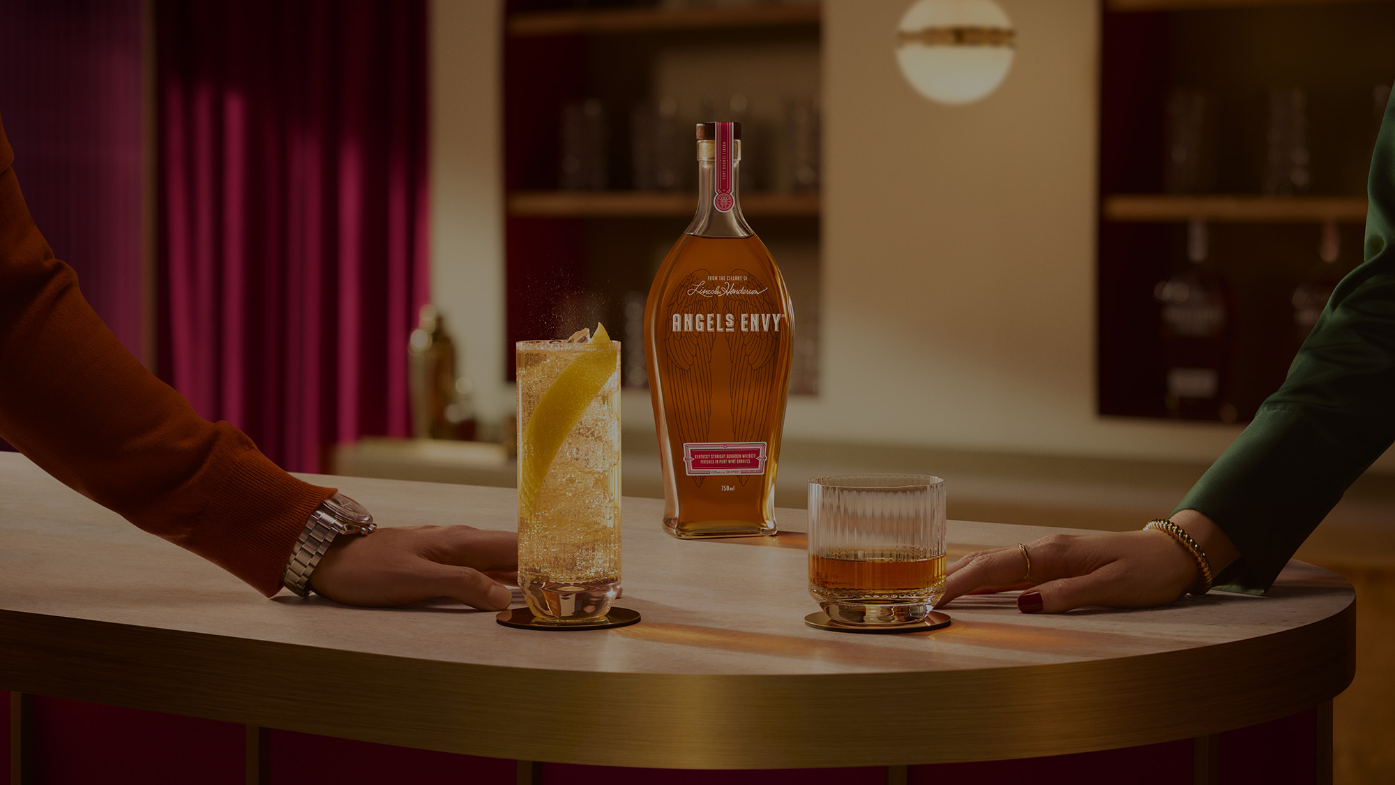 On a white, textured table-top in a modern bar setting with rich ruby design detailing, two hands graze their bourbon cocktails with a bottle of Angel's Envy in the background.