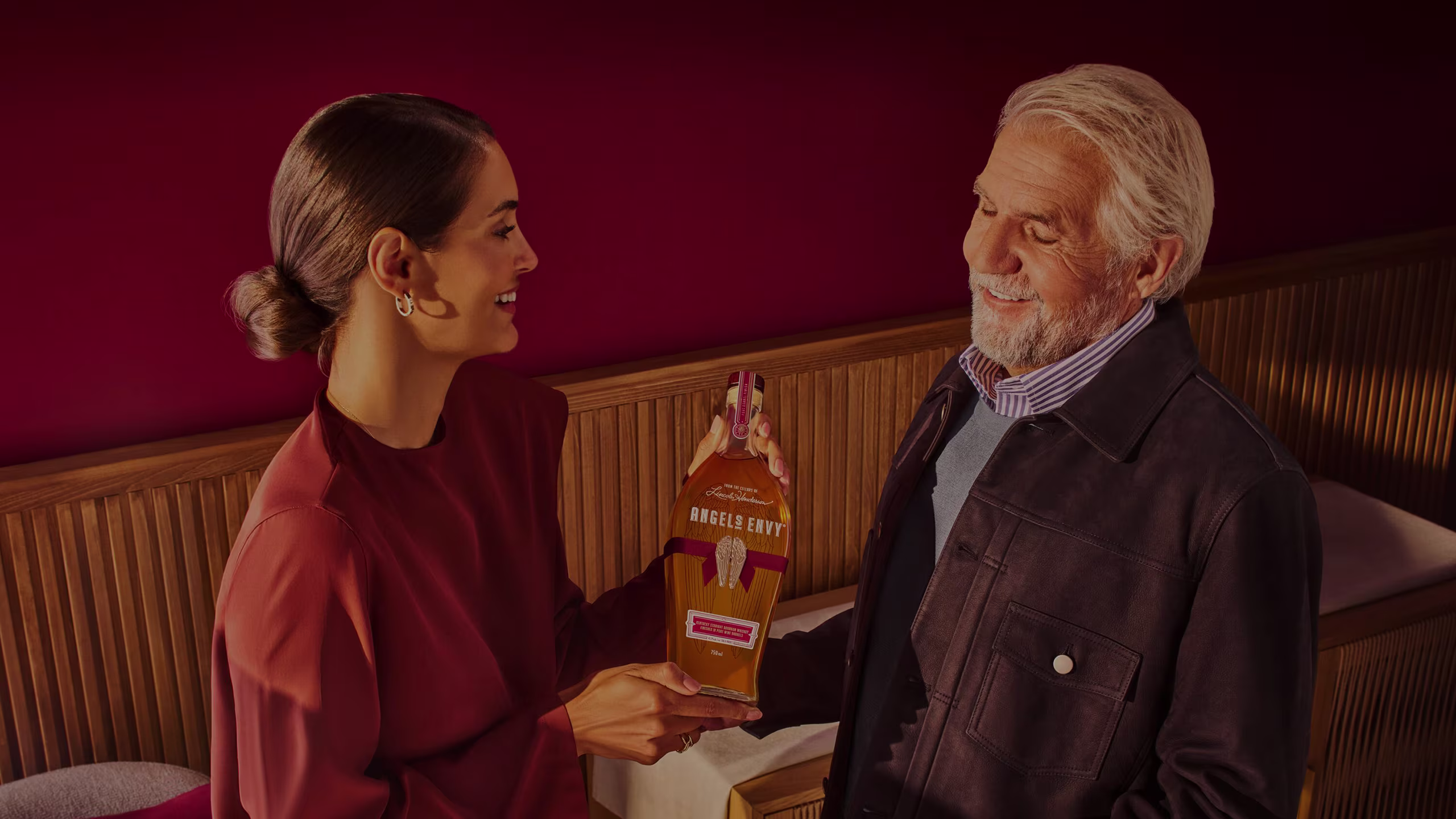 A 30-year old daughter gives a bottle of Angel’s Envy to her older, white-haired father featuring an overlaying text that reads: 2026 Father’s Day Gift Guide.