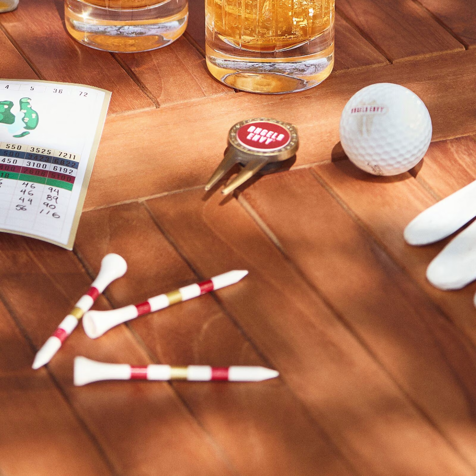 On a wooden outdoor table at the course, Angel’s Envy golf tees, a ball marker, a branded golf ball, a score card and cocktails flicker with sunlight.