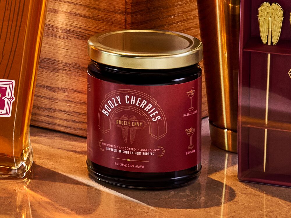 A jar of Boozy Cherries is presented alongside Angel’s Envy bourbon and barware merchandise on a warm wooden bar.