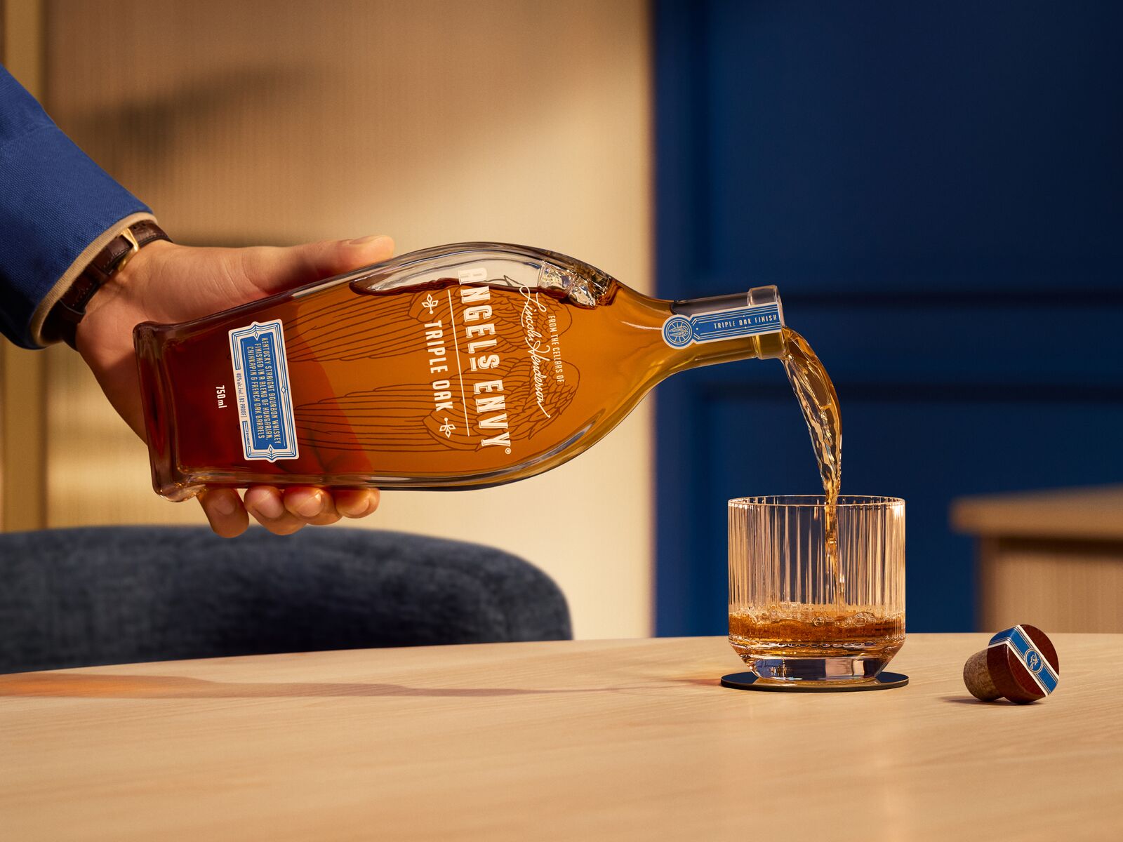A 750 mL bottle of Angel's Envy Triple Oak with a signature wings graphic in black is poured into a rocks glass, set in a modern bar with blue and bright wood design detailing.