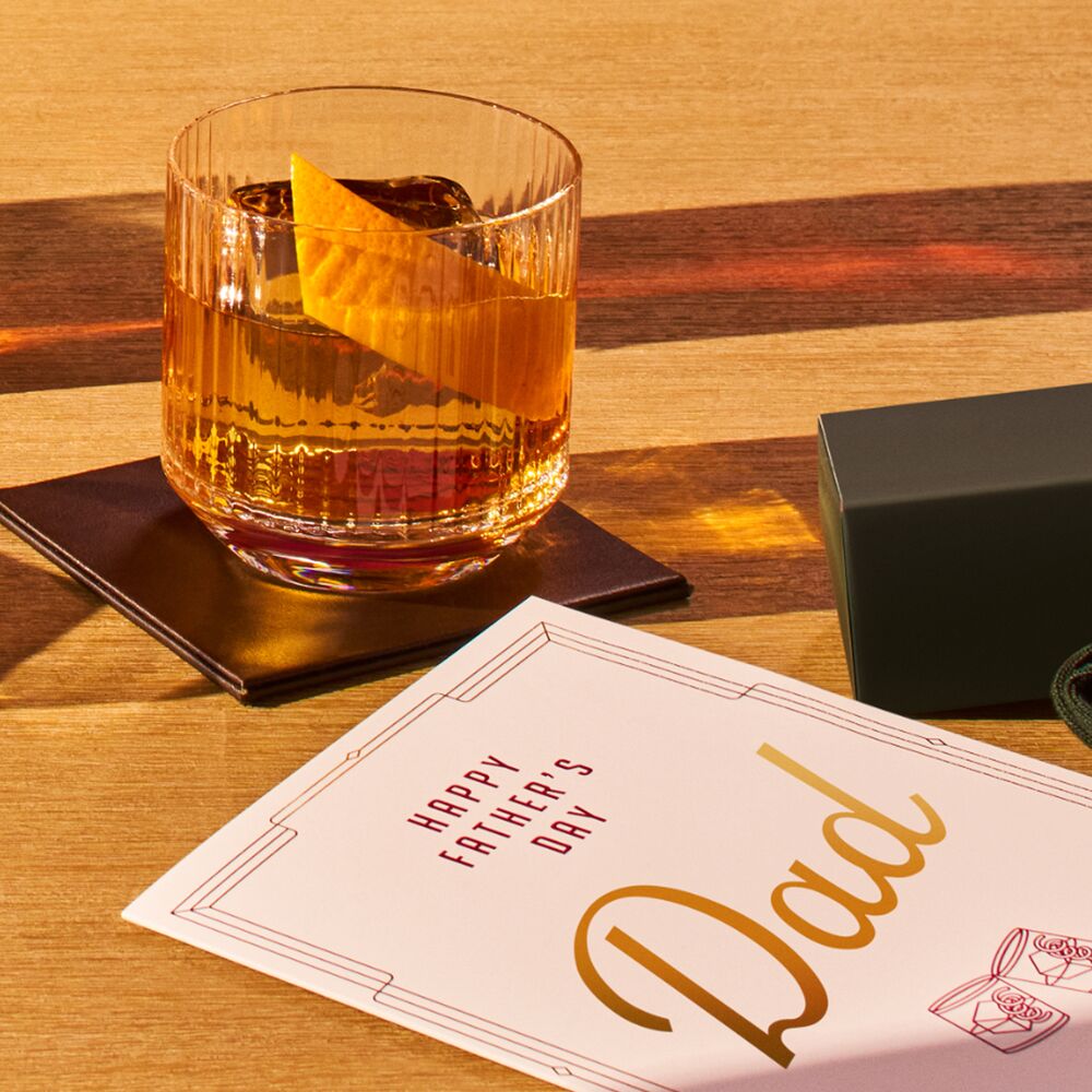 Set on a light wooden table, an Angel’s Envy old fashioned cocktail is presented on a leather coaster beside a Happy Father’s Day card.