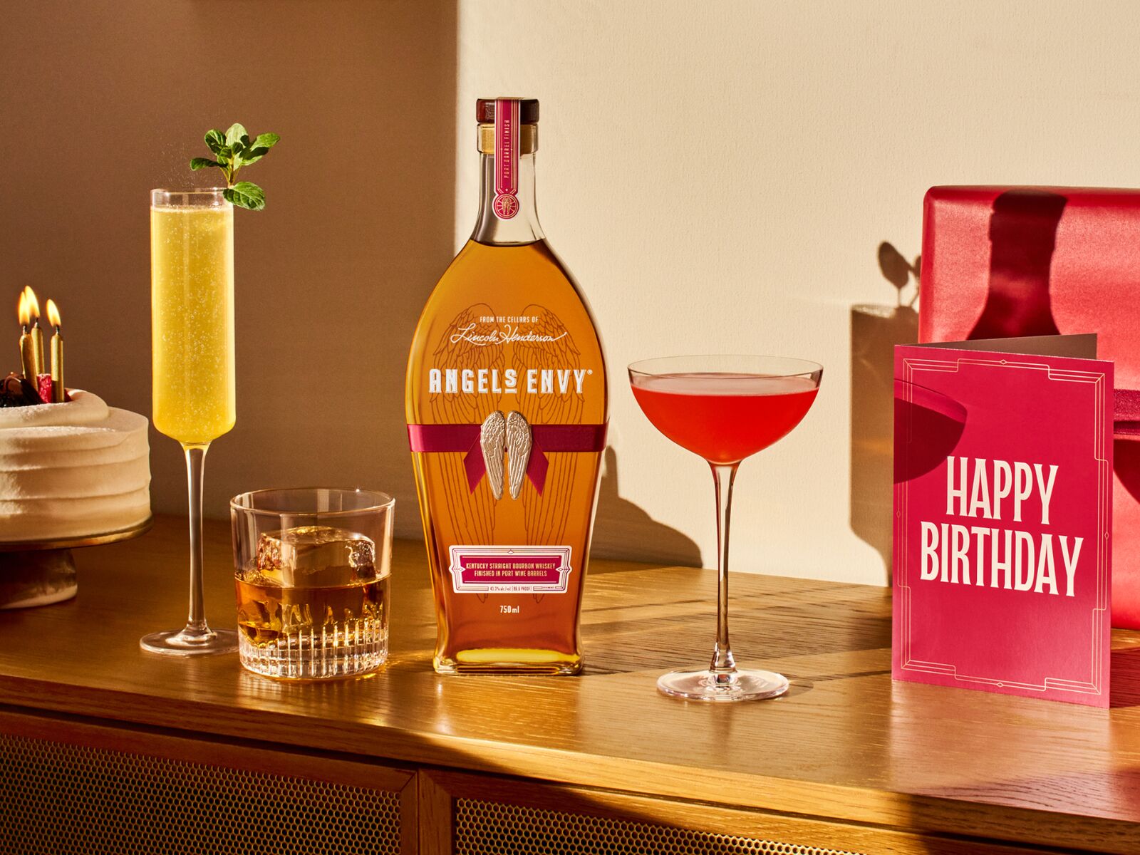 A set of flute, rocks, and coupe glasses accompanied by a bottle of Angel’s Envy and a red card reading “Happy Birthday”.