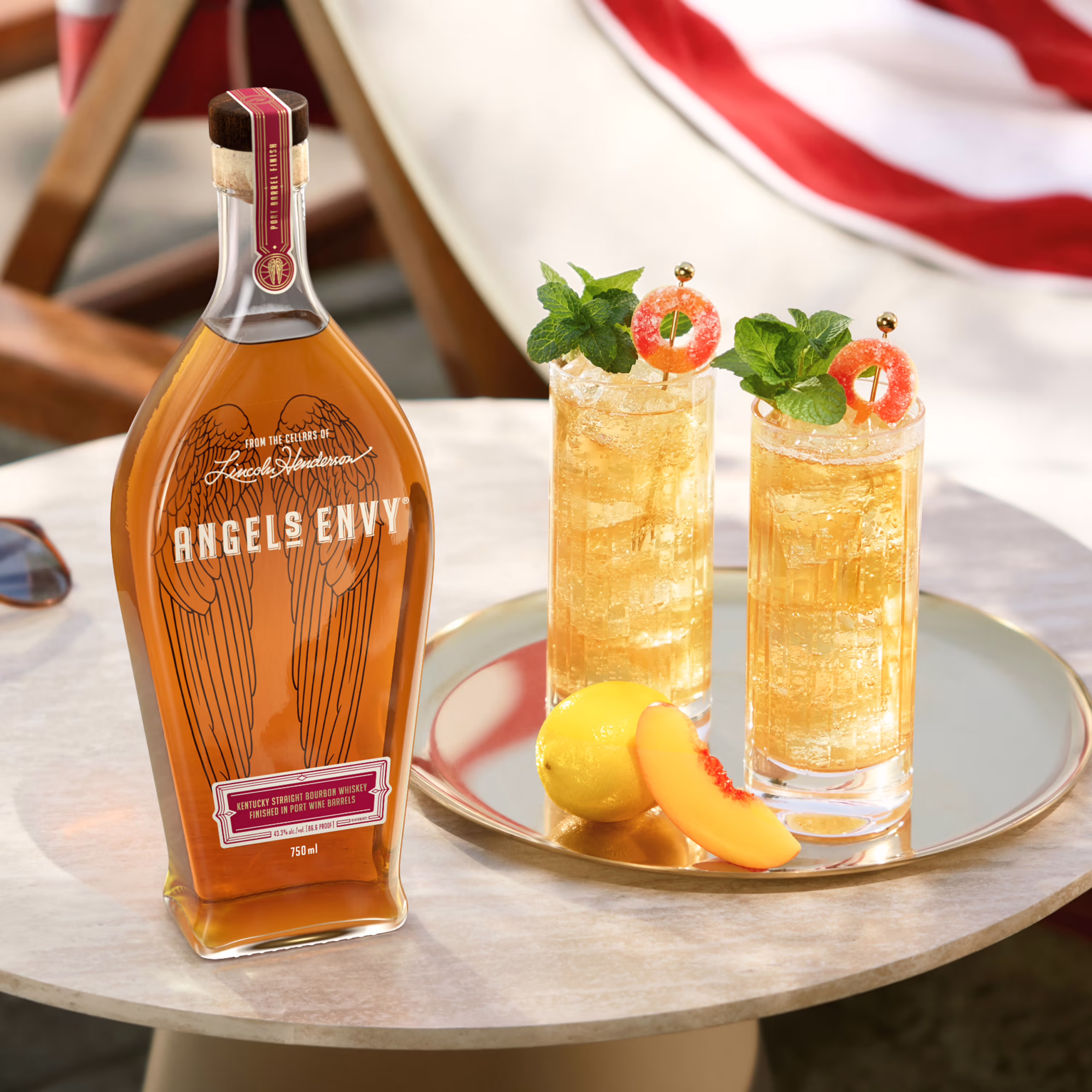 A bottle of Angel’s Envy bourbon next to two ice-cold cocktails, served atop a sunlit poolside table on a relaxing summer day. They’re styled with fresh fruit and garnished with mint and sour candy.